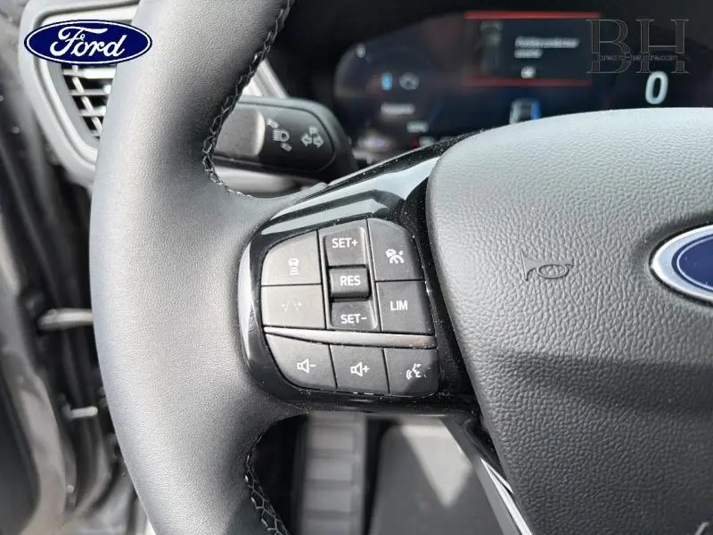 Gros plan sur les commandes au volant noir du Ford Kuga 2026 hybride, avec logo Ford visible au centre.