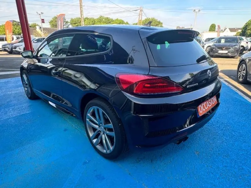 Vue 3/4 arrière droite d'une Volkswagen Scirocco noire intense avec jantes alu et becquet arrière sur parking.