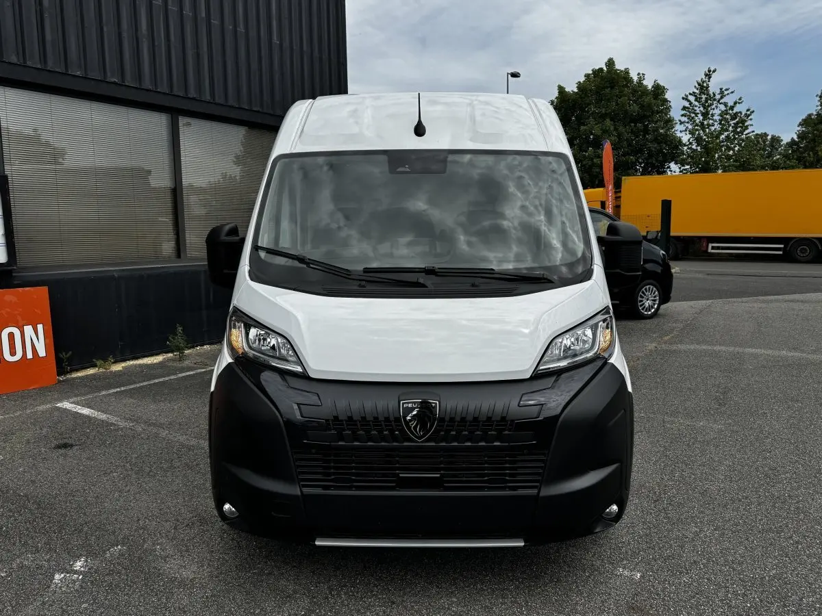 Vue avant d’un Peugeot Boxer Fourgon blanc avec calandre noire et phares allumés sur un parking extérieur.