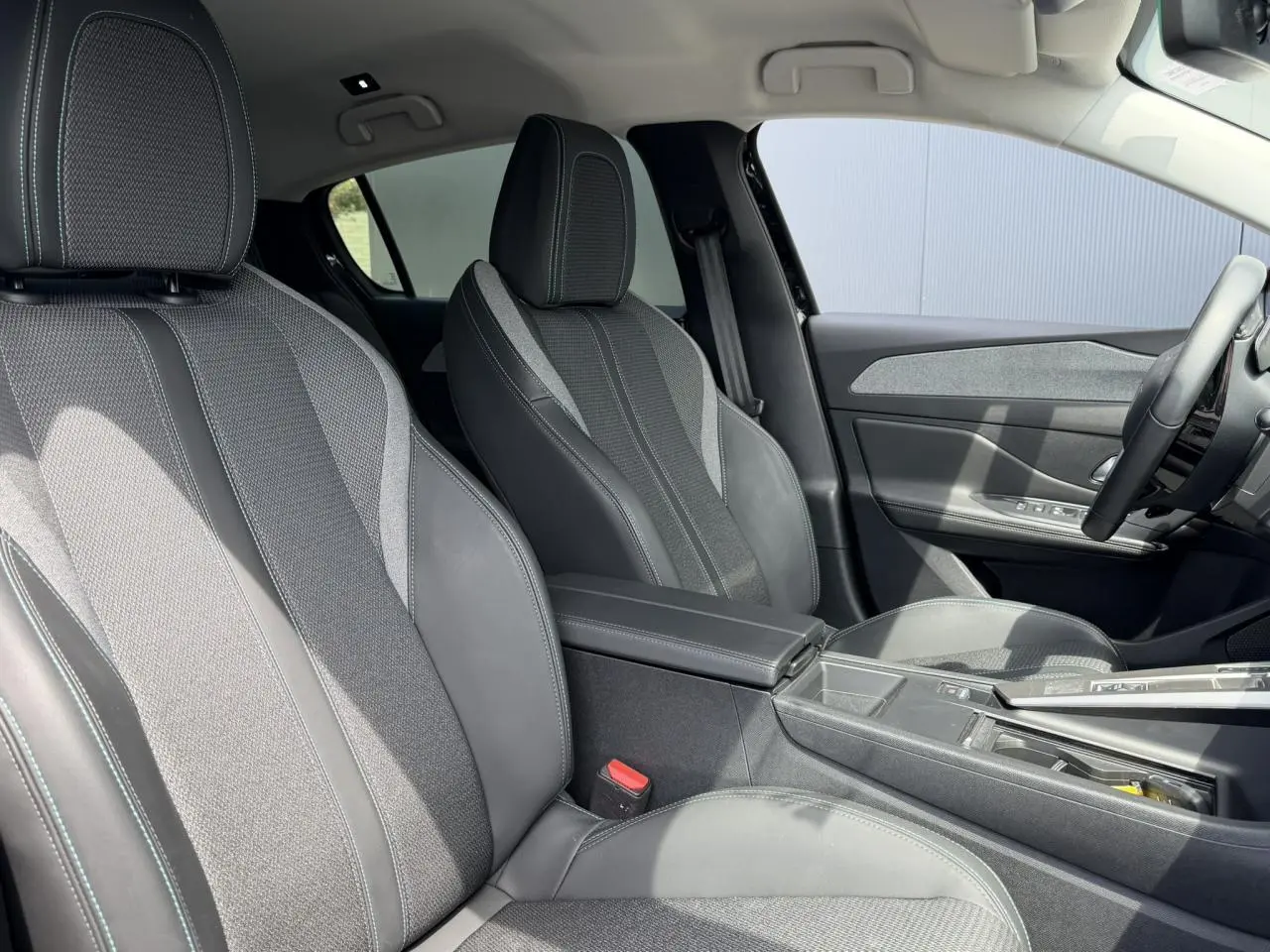 Vue intérieure côté gauche des sièges avant gris foncé avec surpiqûres bleues et accoudoir central de la Peugeot 308 Hybrid 2025.
