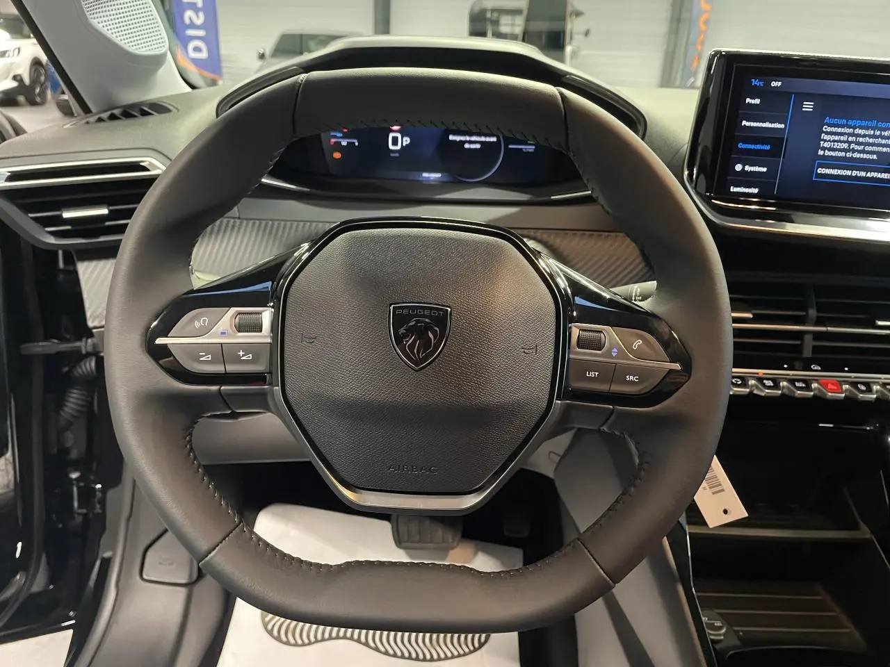 Vue rapprochée du volant cuir noir de la Peugeot 208 1.2i Hybrid avec tableau de bord numérique et commandes intégrées.