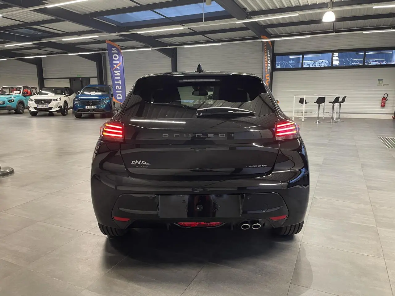 Vue arrière d'une Peugeot 208 noire métallisée hybride, avec feux arrière allumés et double sortie d'échappement chrome.