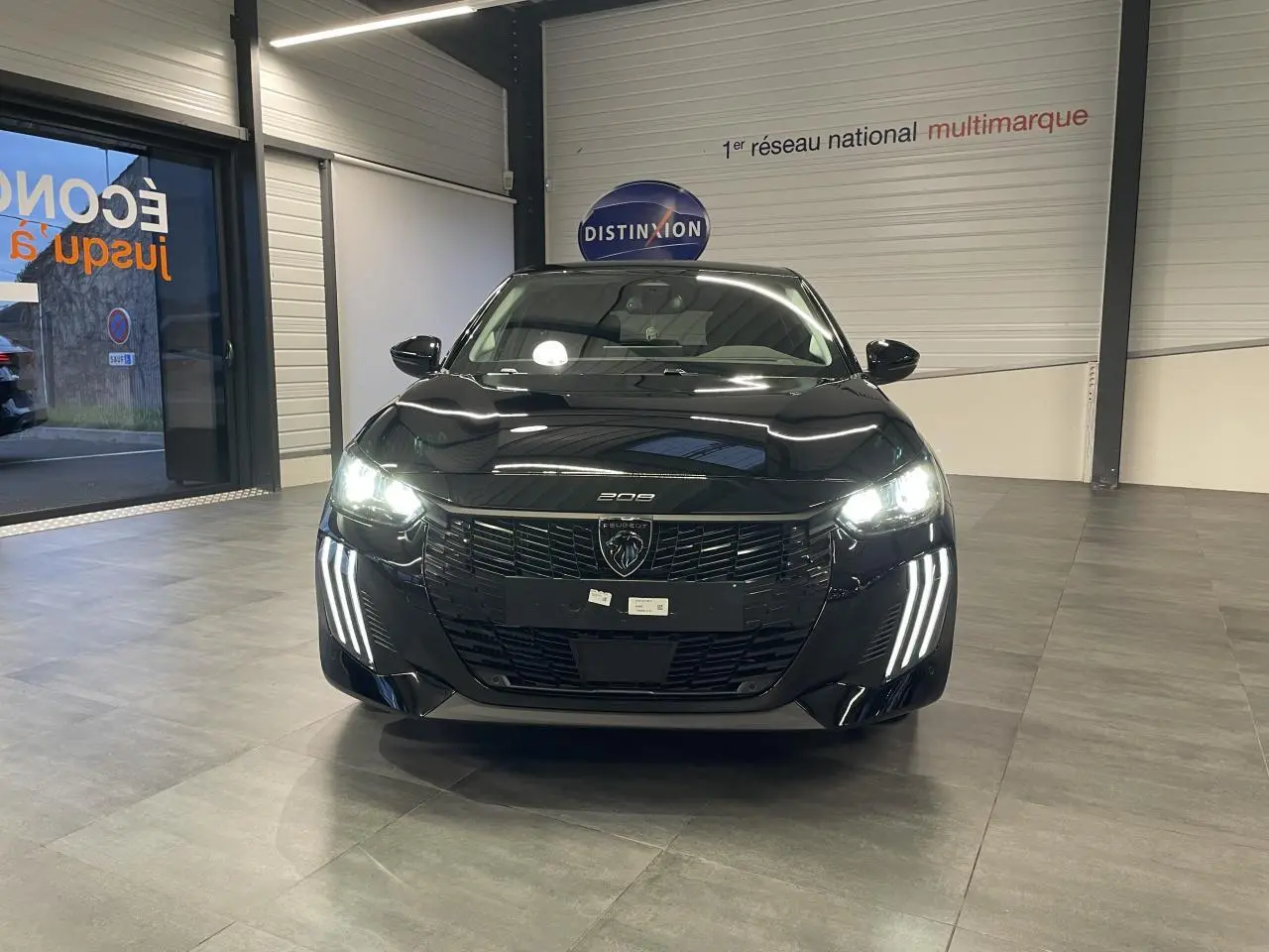 Vue avant d'une Peugeot 208 noire métallisée avec phares et feux diurnes LED allumés en intérieur showroom.