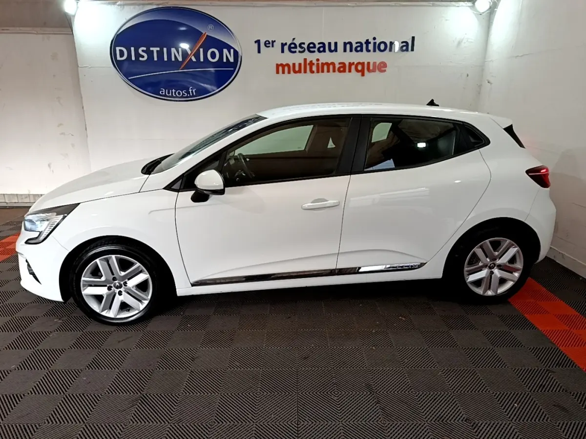 Profil côté gauche d'une Renault Clio Business TCe 100 blanche, avec jantes alliage et feux arrière allumés, en intérieur showroom.
