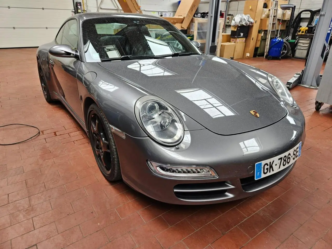 Porsche 911 Carrera 4S gris météore vue 3/4 avant gauche dans un atelier avec jantes noires et phares ronds.
