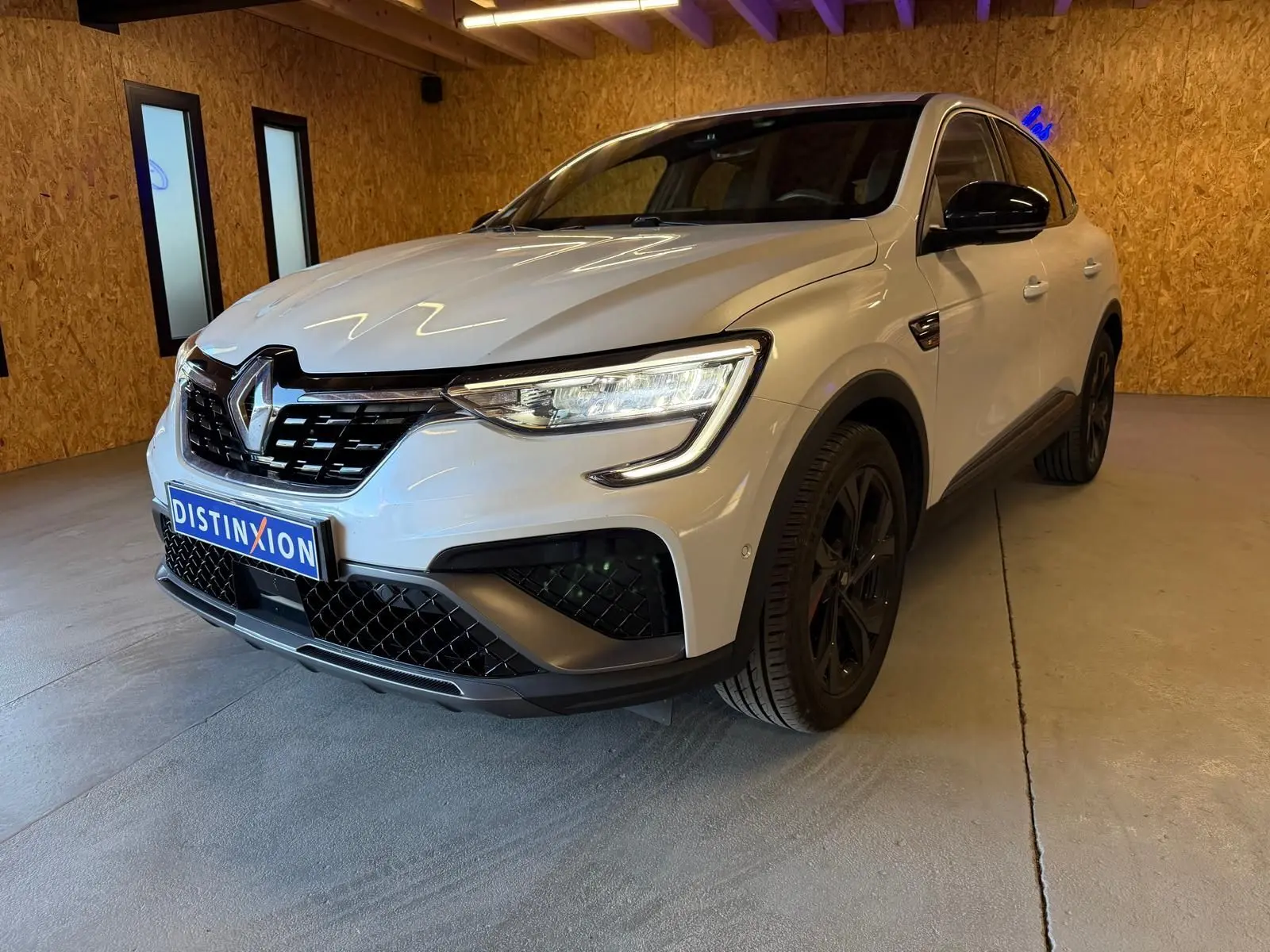 Renault Arkana blanc nacré en 3/4 avant droit, avec calandre noire et jantes alu noires, stationné en intérieur.