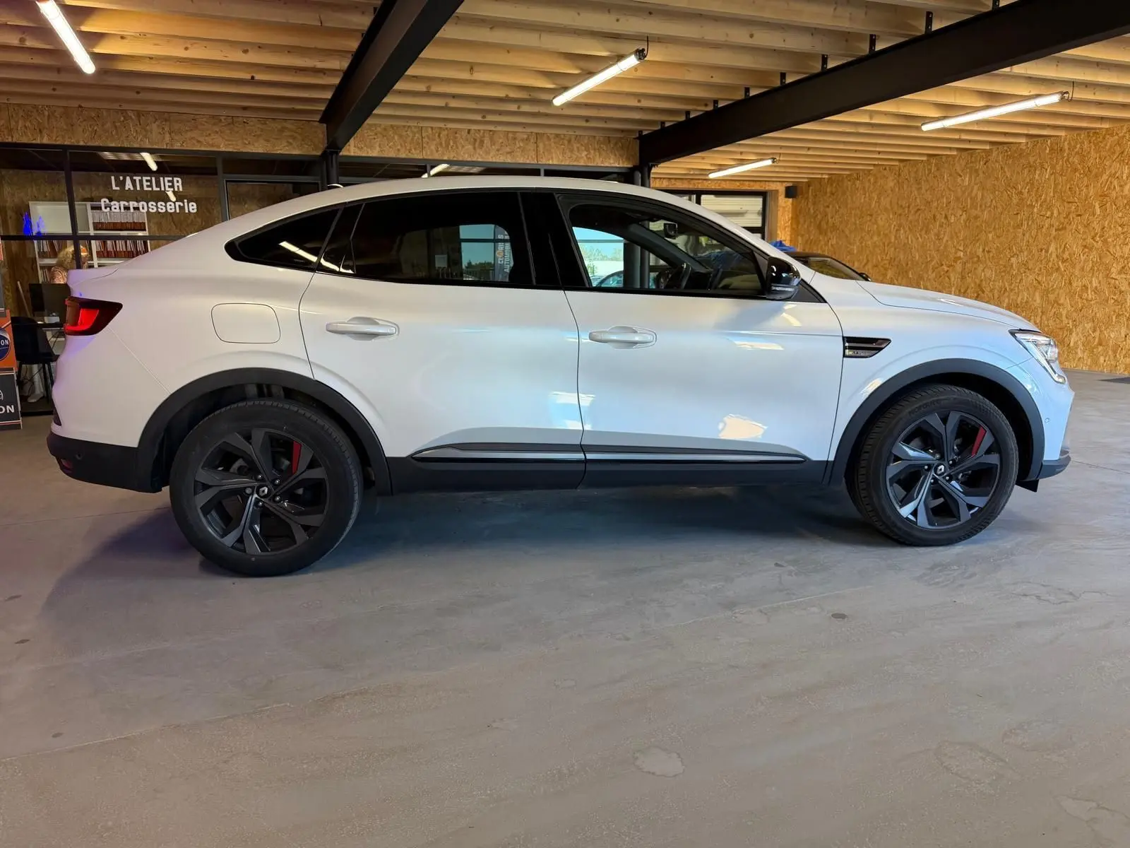 Vue de profil côté gauche d'un Renault Arkana blanc nacré avec jantes alu noires et étriers rouges, stationné en intérieur.