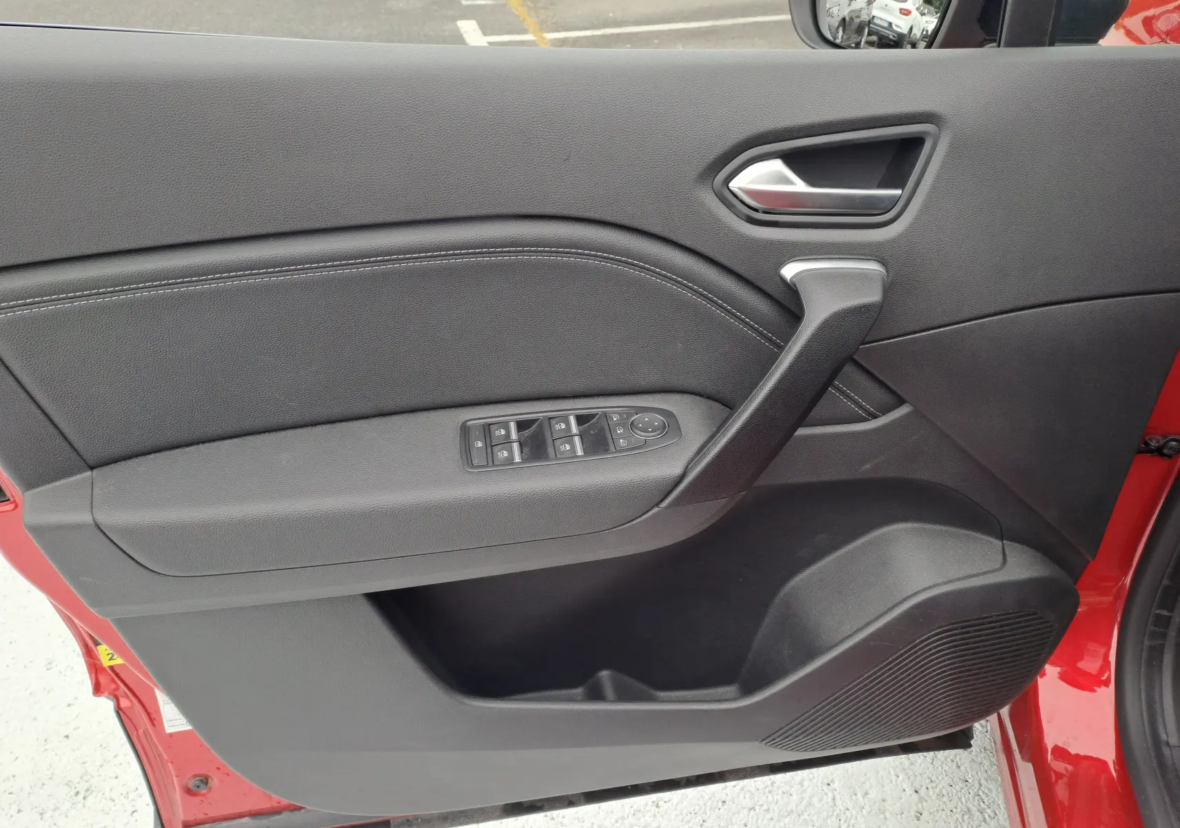 Intérieur côté gauche de la porte avant d'un Renault Captur rouge flamme métal, avec commandes électriques et poignée noire.
