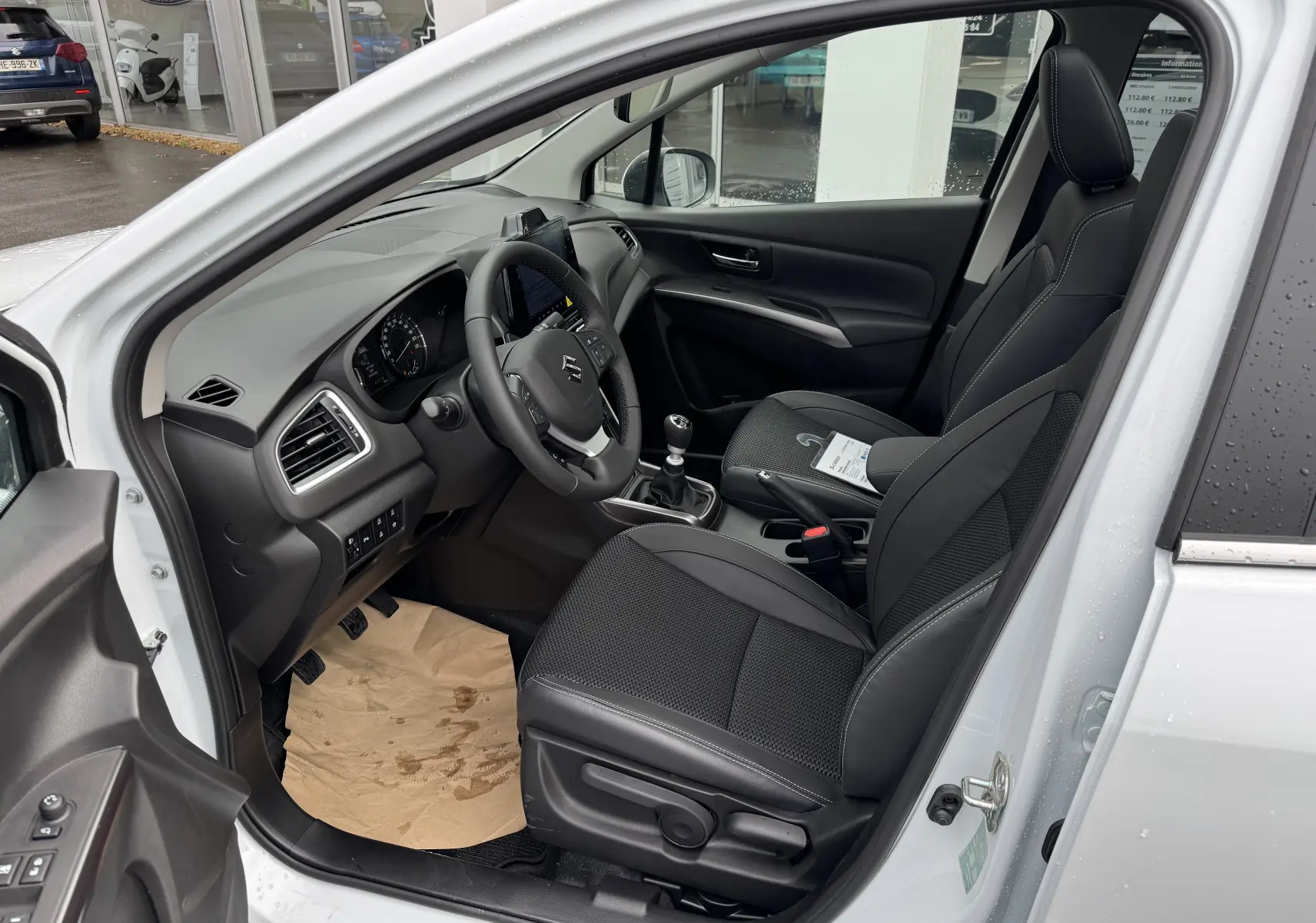 Vue intérieure côté conducteur du Suzuki S-Cross blanc 2025, mettant en valeur les sièges noirs et la boîte manuelle.