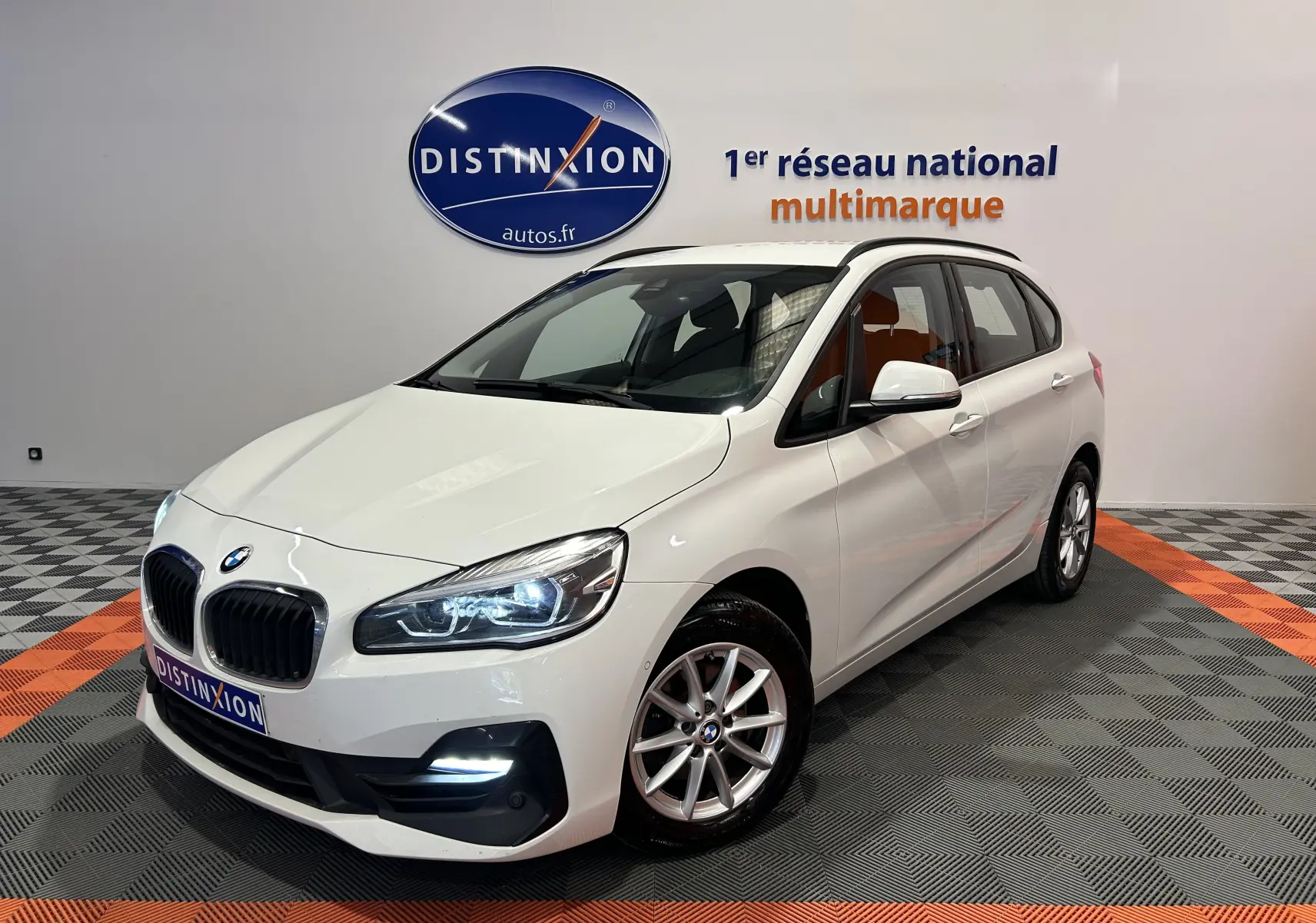 BMW Série 2 Active Tourer 216d blanc vue 3/4 avant droit avec phares allumés dans showroom.