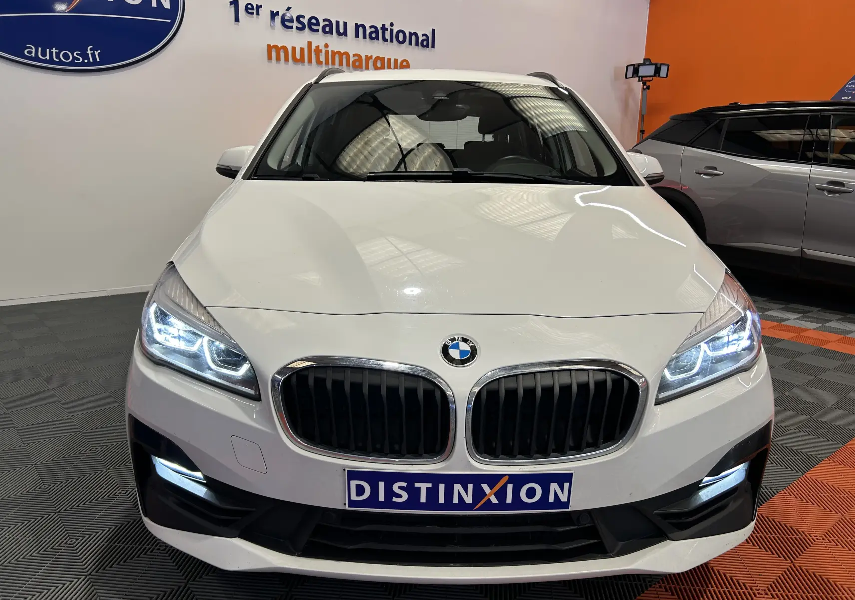 Vue frontale d'une BMW Série 2 Active Tourer 216d blanche avec phares LED allumés dans un showroom.