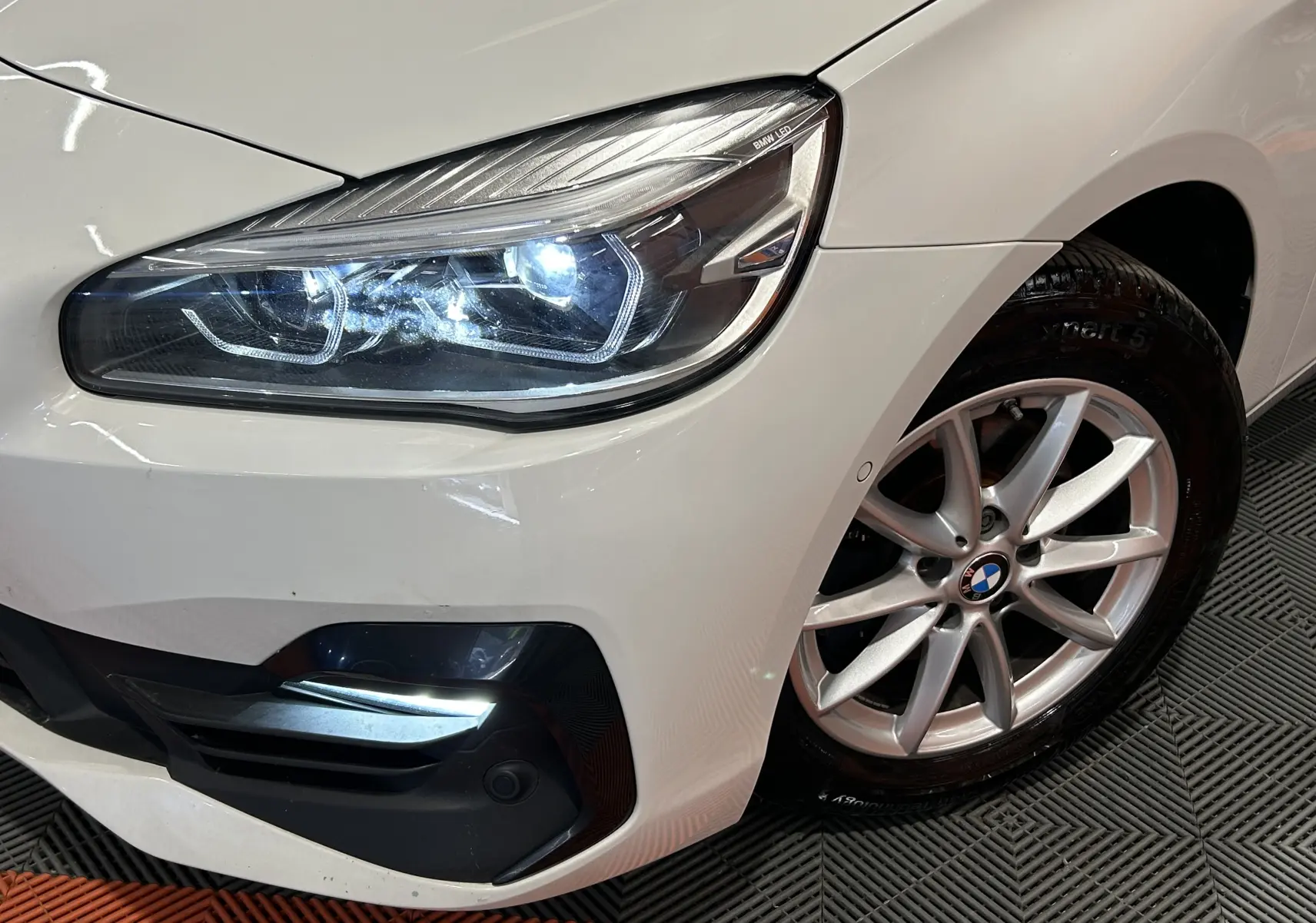 Gros plan sur l’avant droit blanc d’une BMW Série 2 Active Tourer 216d avec phares LED allumés et jante alliage.