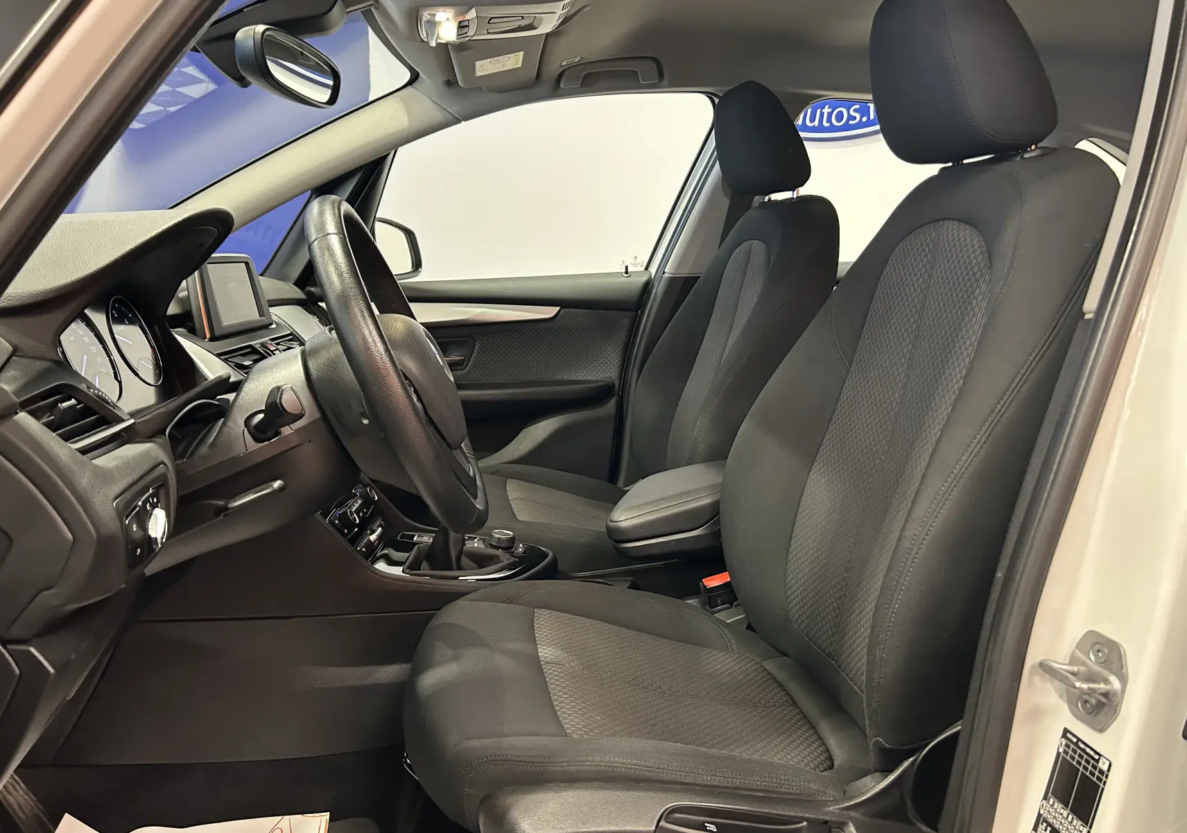 Vue intérieure côté conducteur de la BMW Série 2 Active Tourer blanche, montrant les sièges en tissu noir avec appuie-têtes et volant multifonction.