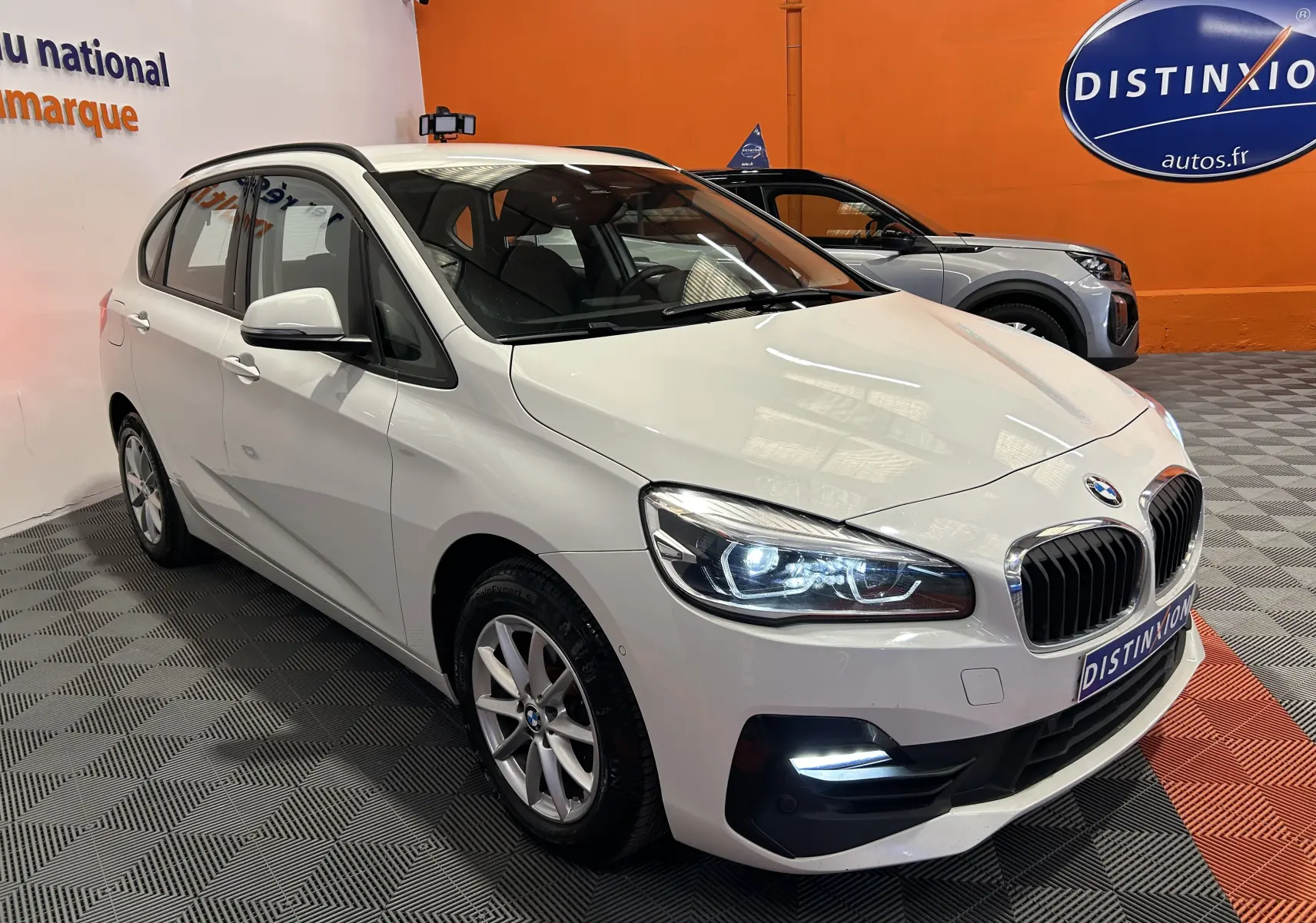 BMW Série 2 Active Tourer 216d blanc vue 3/4 avant droit avec phares et feux diurnes allumés dans showroom.