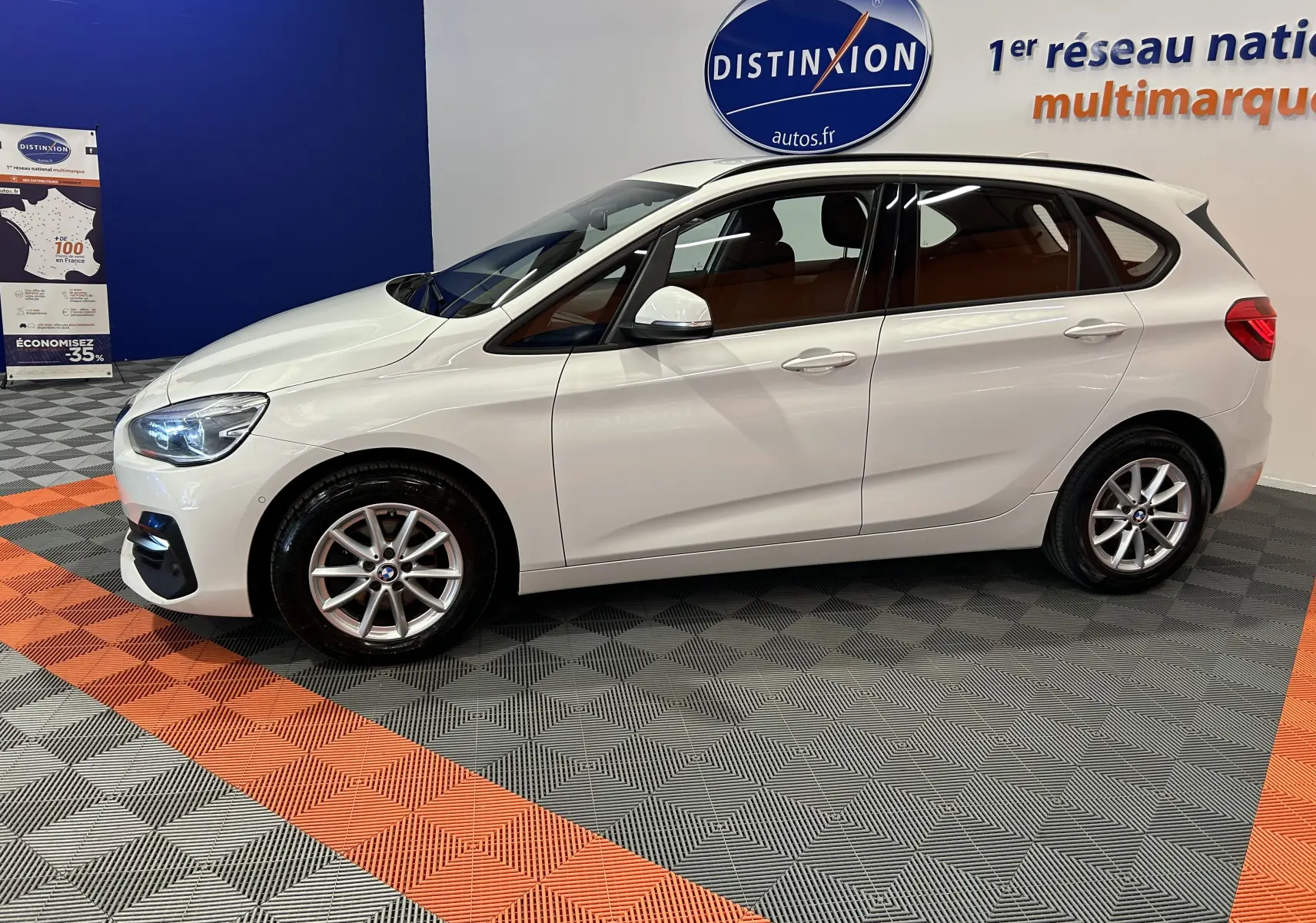 BMW Série 2 Active Tourer 216d blanc vue de profil côté gauche en intérieur, avec jantes alliage et feux allumés.