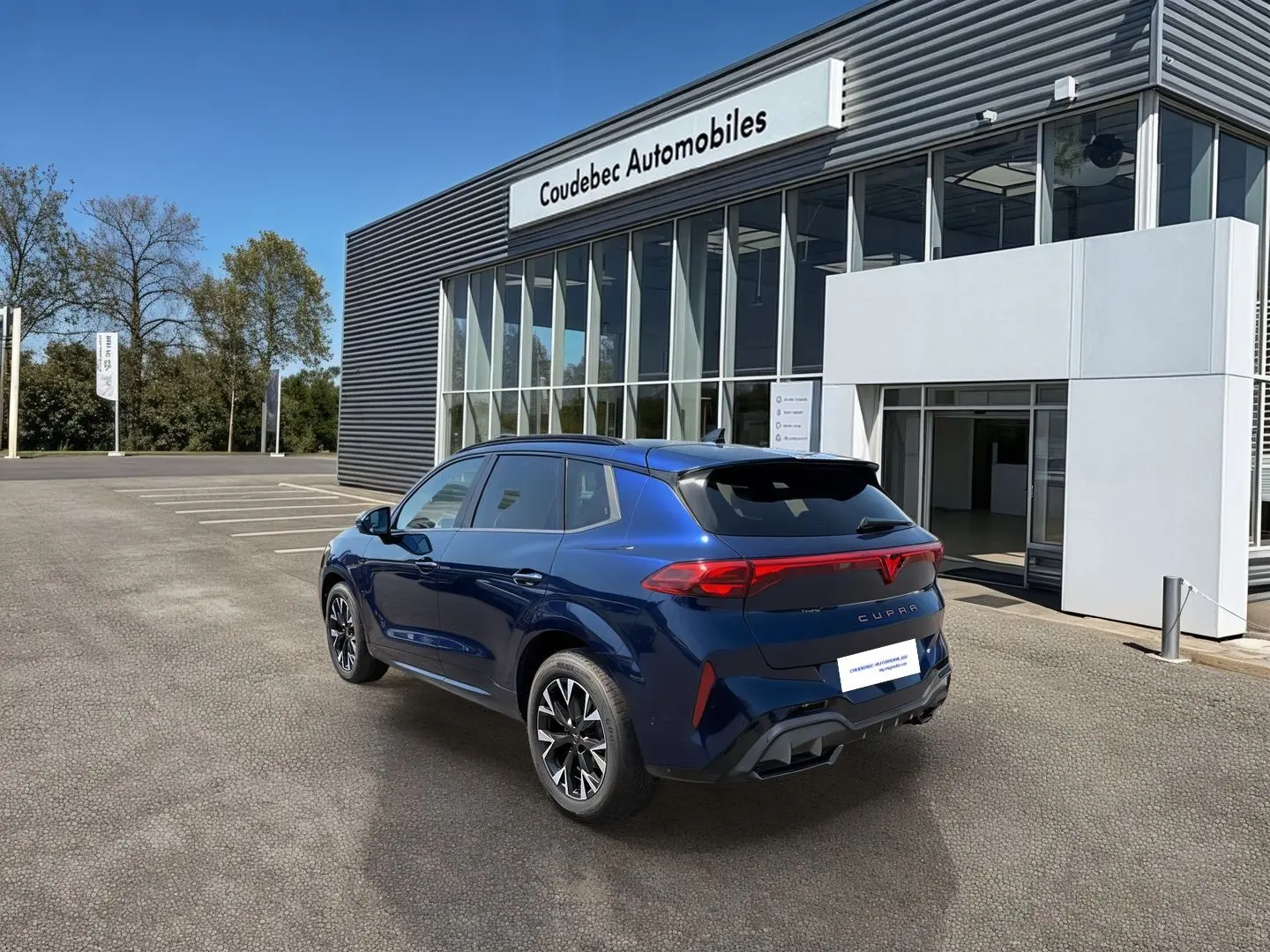 Vue 3/4 arrière droite d'un CUPRA Terramar bleu métallisé devant un bâtiment moderne avec logo CUPRA lumineux à l'arrière.