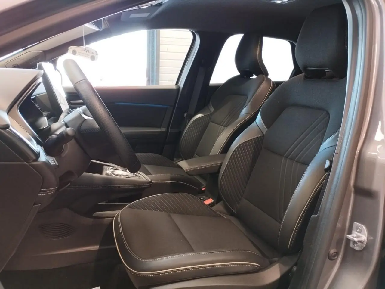 Vue intérieure côté conducteur de la Renault Symbioz 2025 gris Cassiopée, sièges noirs avec surpiqûres claires et console centrale.