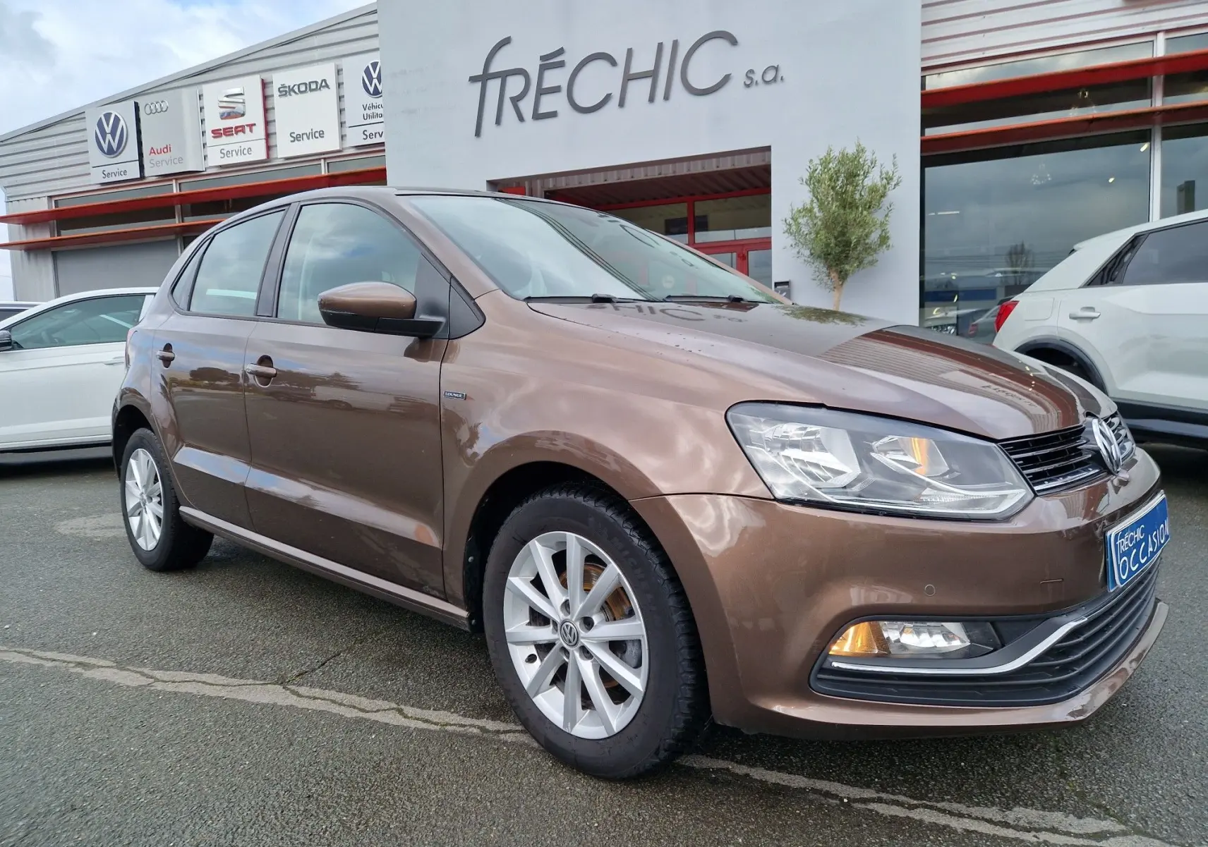 Volkswagen Polo 1.4 TDI brun vue 3/4 avant droit, avec jantes alliage et feux allumés devant un concessionnaire.