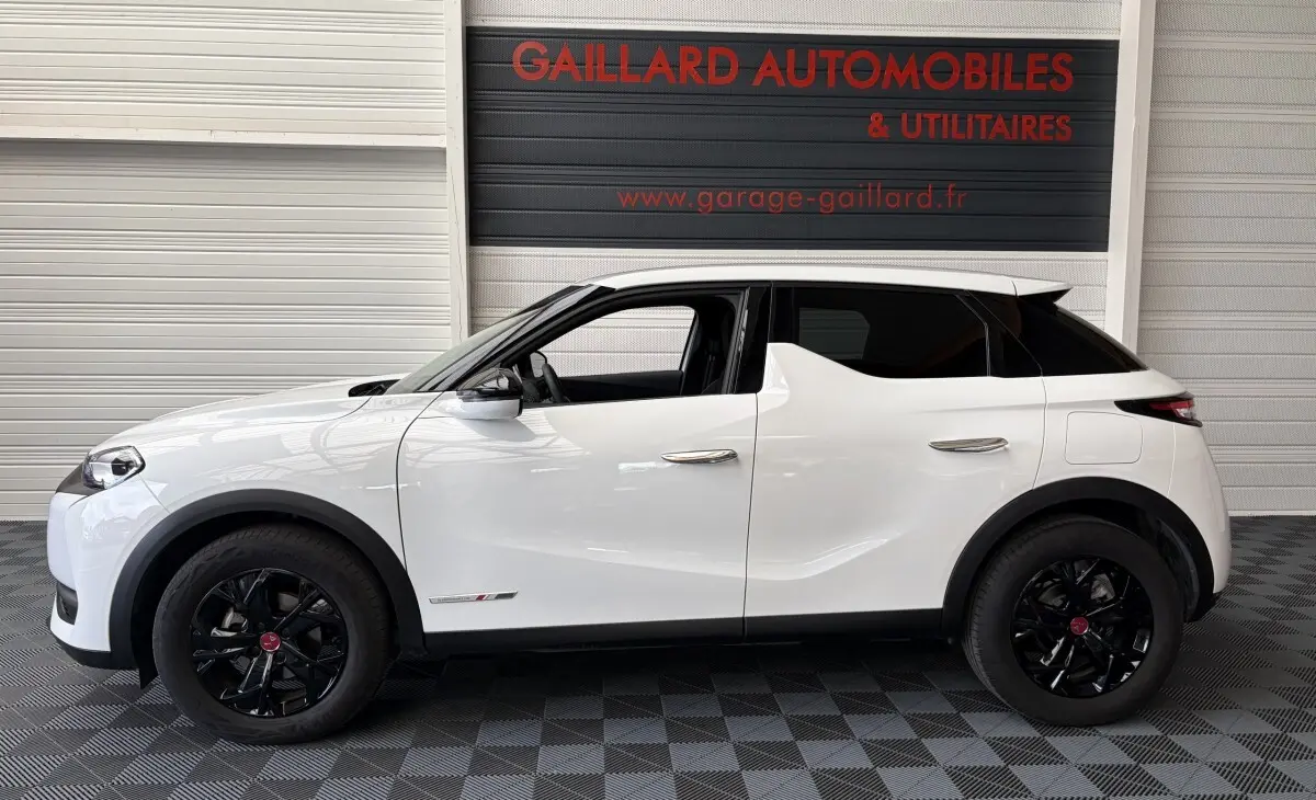 Vue latérale droite d'un DS3 Crossback blanc avec jantes noires et vitres arrière teintées, en intérieur showroom.