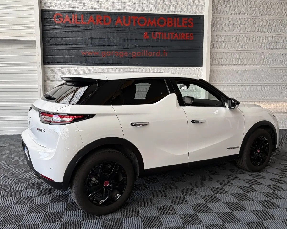 Vue 3/4 arrière droite d'un DS3 Crossback blanc avec toit noir et jantes noires dans un garage.
