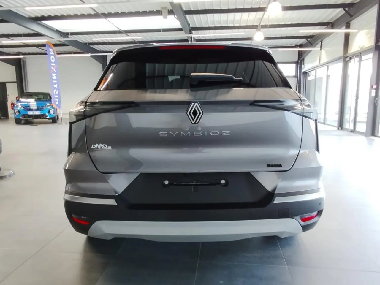 Vue arrière du Renault Symbioz gris Cassiopée avec logo central et feux arrière noirs intégrés, en intérieur showroom.