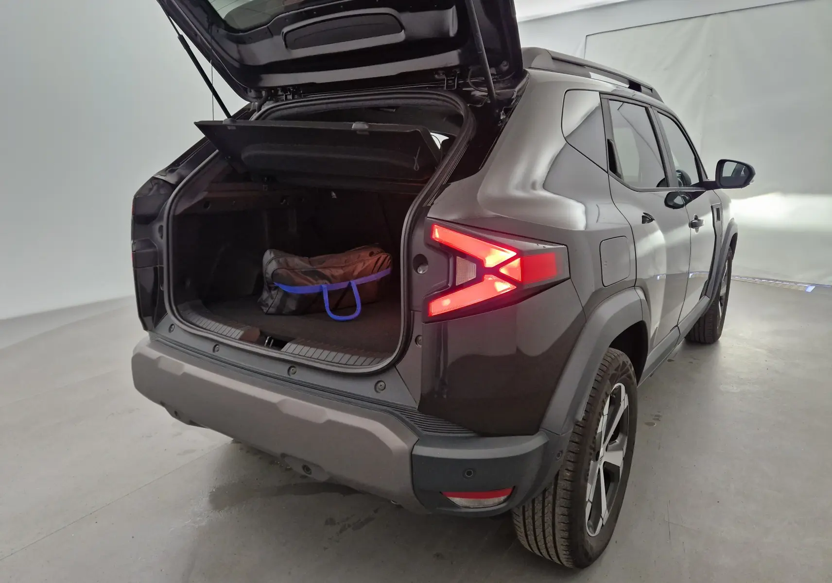Vue 3/4 arrière droite du Dacia Duster noir 2024 avec coffre ouvert et sac à l'intérieur, feux arrière allumés.