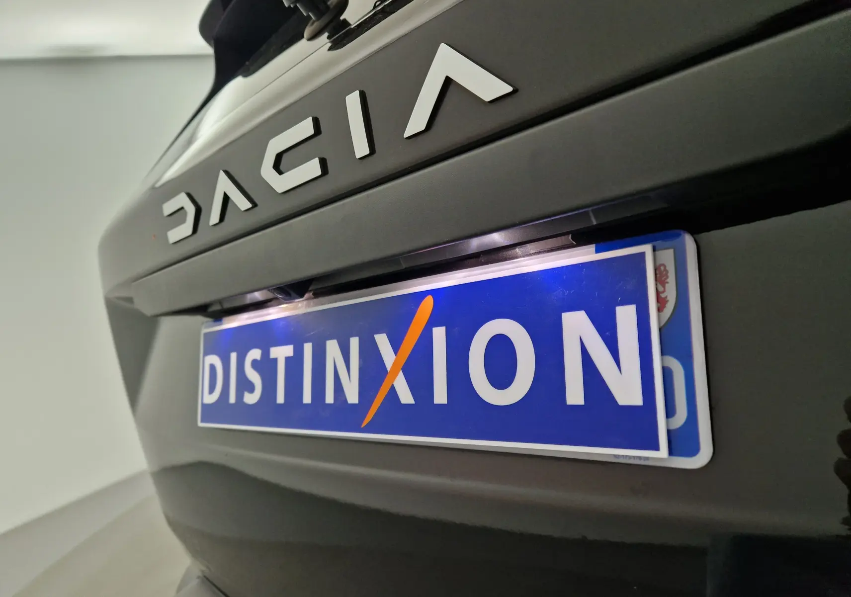 Gros plan sur le logo DACIA et la plaque avant bleue "DISTINXION" d'un Duster noir 2024 en intérieur.