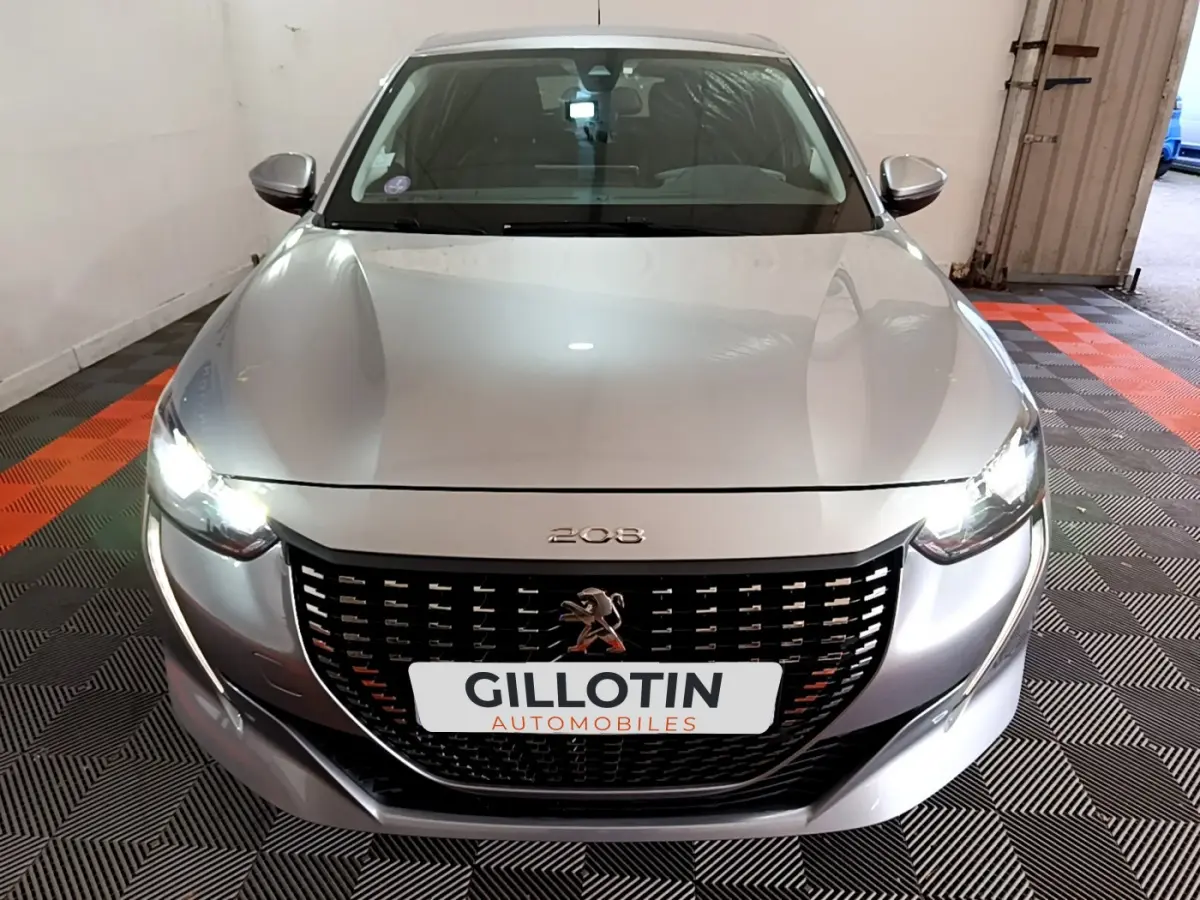 Vue avant d'une Peugeot 208 gris clair avec phares LED allumés dans un garage fermé.