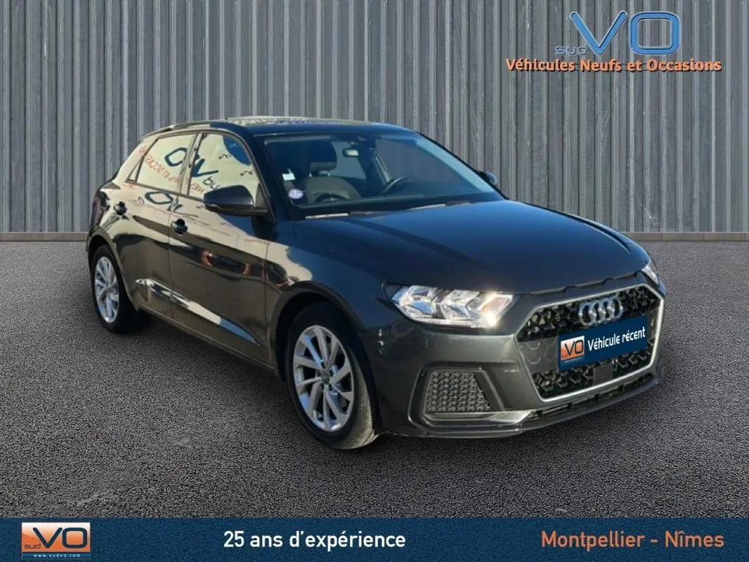 Audi A1 Sportback gris foncé vue 3/4 avant droit avec phares allumés sur fond urbain neutre.