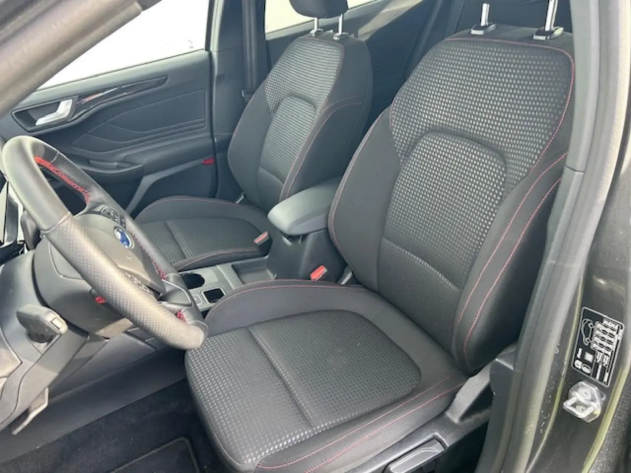 Vue intérieure côté conducteur de la Ford Focus IV ST-Line X, sièges tissu noir avec surpiqûres rouges et volant sport.