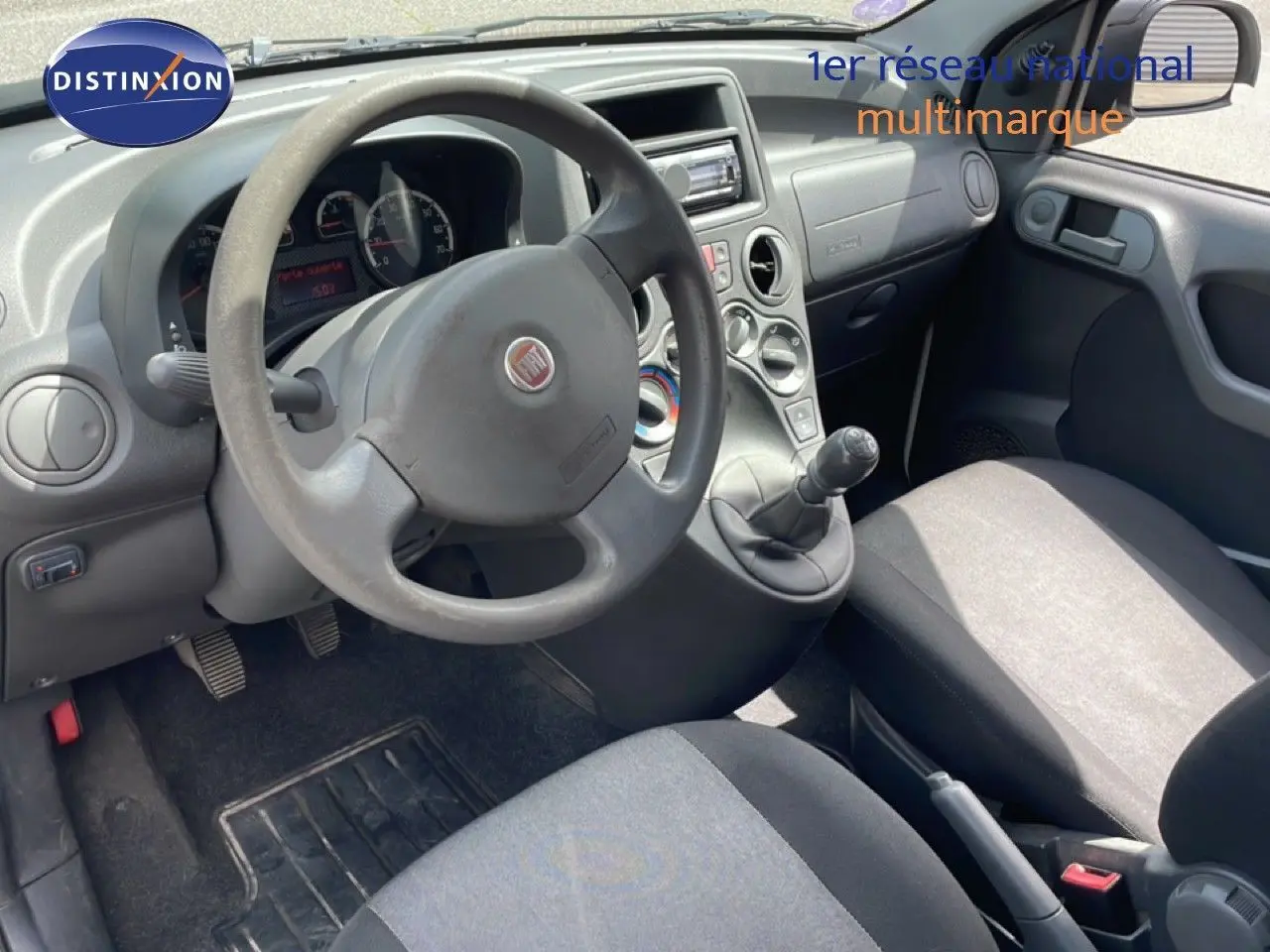 Vue intérieure côté conducteur de la Fiat Panda blanche 2011, volant, tableau de bord et sièges en tissu gris visibles.
