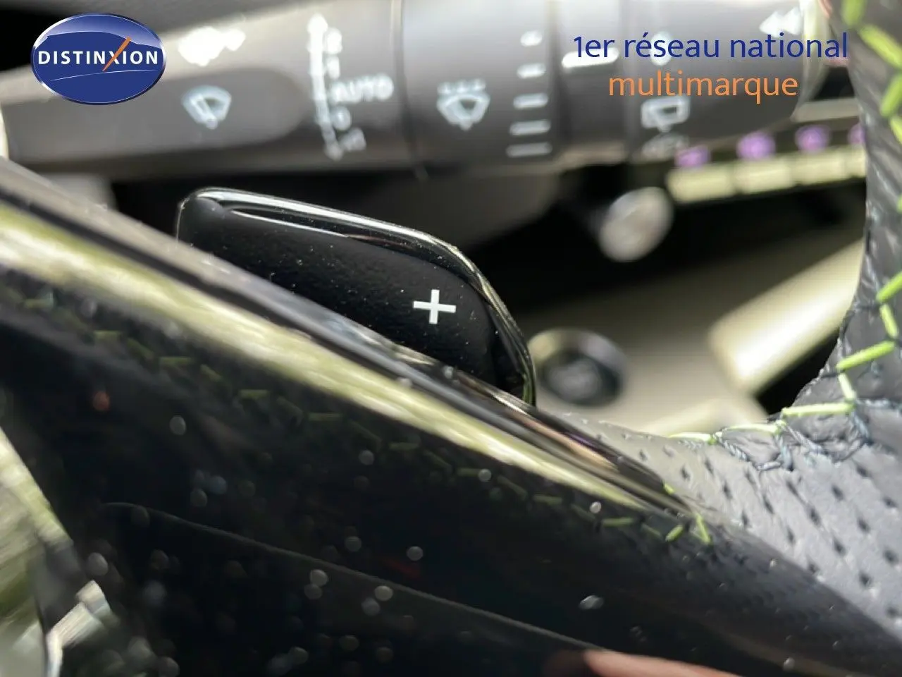 Détail du levier de commande + sur le volant en cuir noir avec surpiqûres vertes d'une Peugeot 308 hybride 2026.