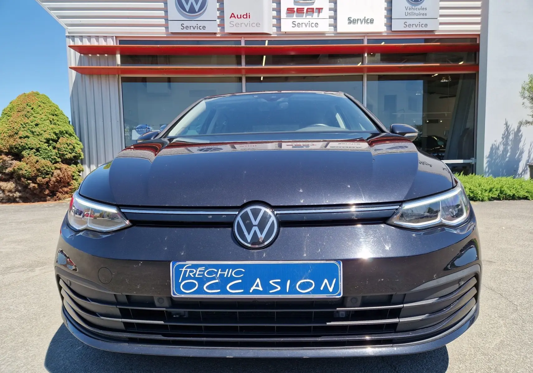 Vue frontale d'une Volkswagen Golf VIII noire métallisée garée devant un concessionnaire multimarques.