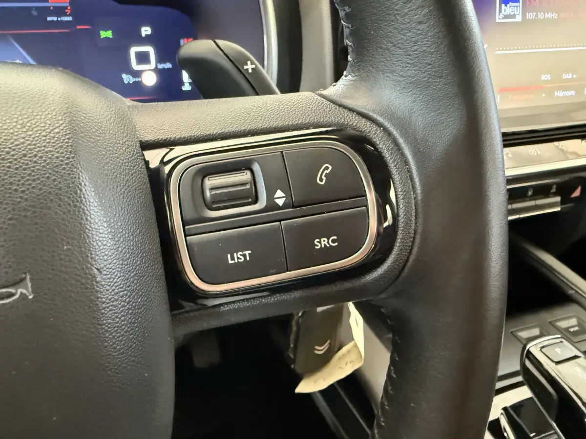 Gros plan sur les commandes gauche du volant cuir noir du Citroën C5 Aircross gris clair, avec écran tactile en arrière-plan.