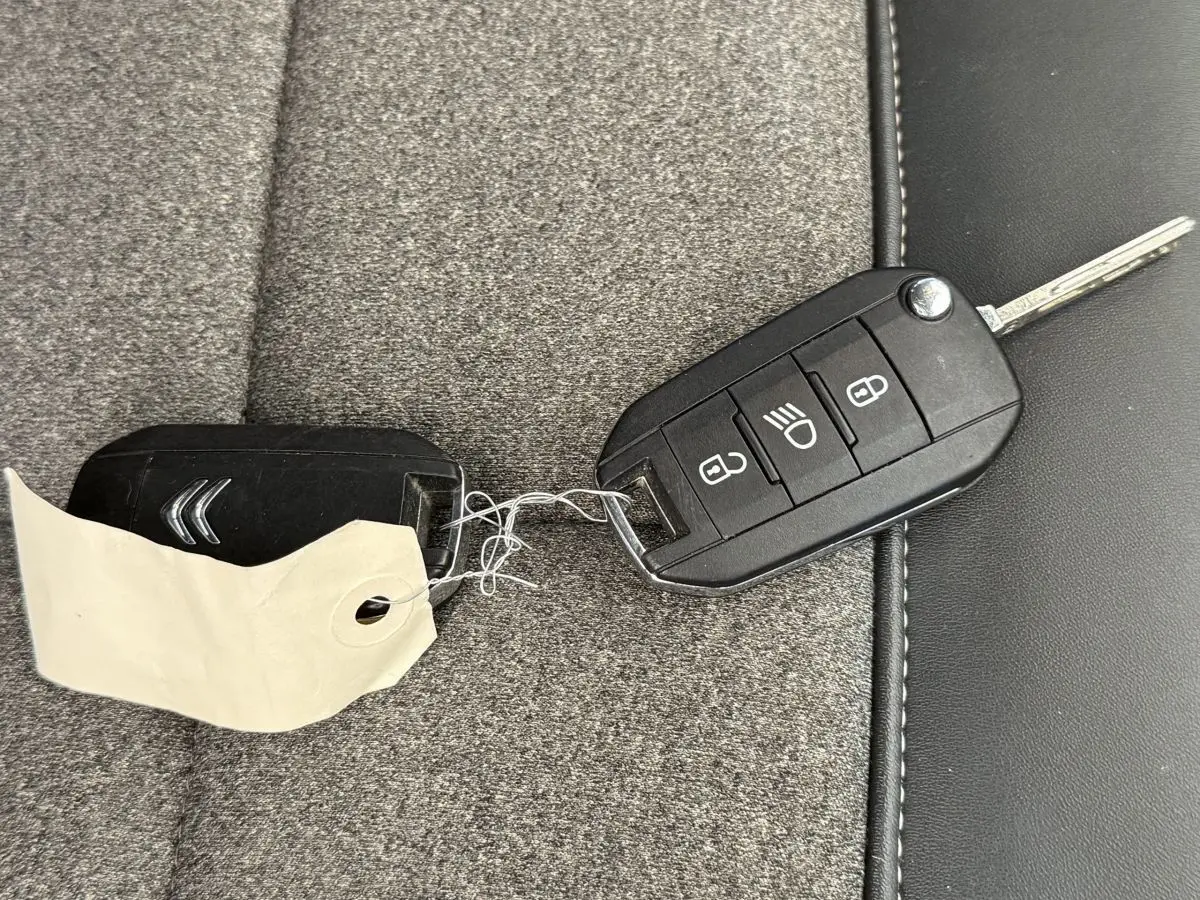 Clé de voiture noire Citroën posée sur siège tissu gris et cuir noir, avec porte-clés et étiquette blanche.