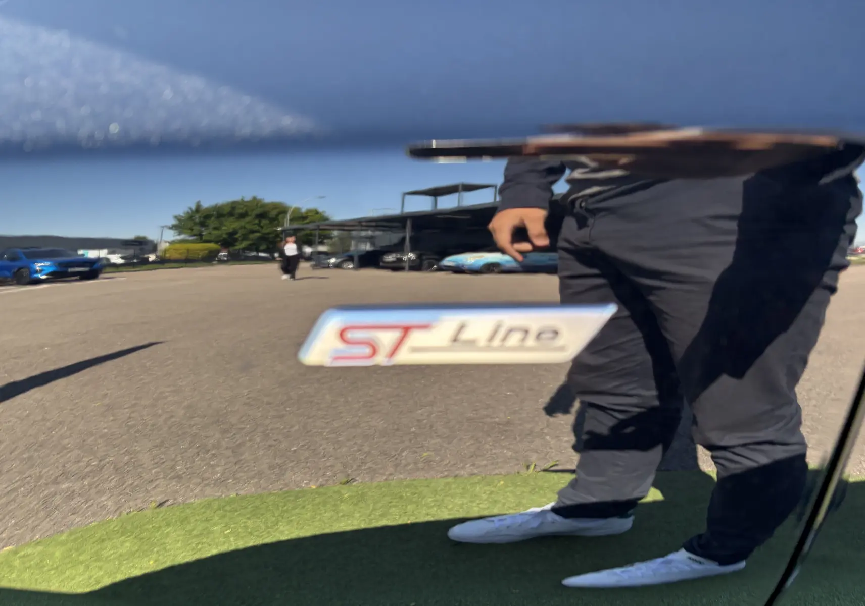 Gros plan sur le badge ST-Line sur la portière noire Agate d'une Ford Focus IV, reflet d'une personne et du parking visible.
