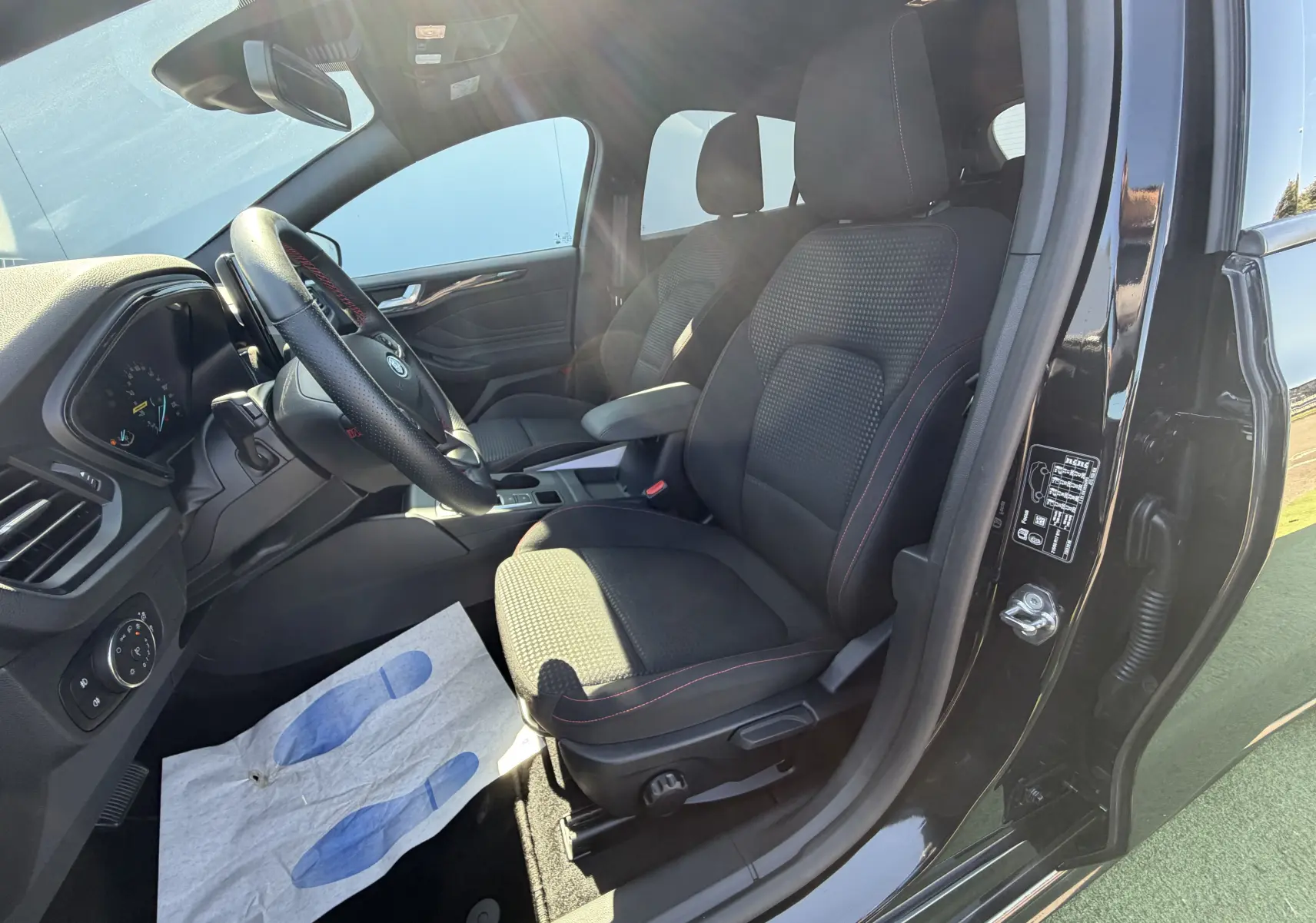 Vue intérieure côté conducteur de la Ford Focus IV ST-Line noire avec sièges tissu et surpiqûres rouges visibles.