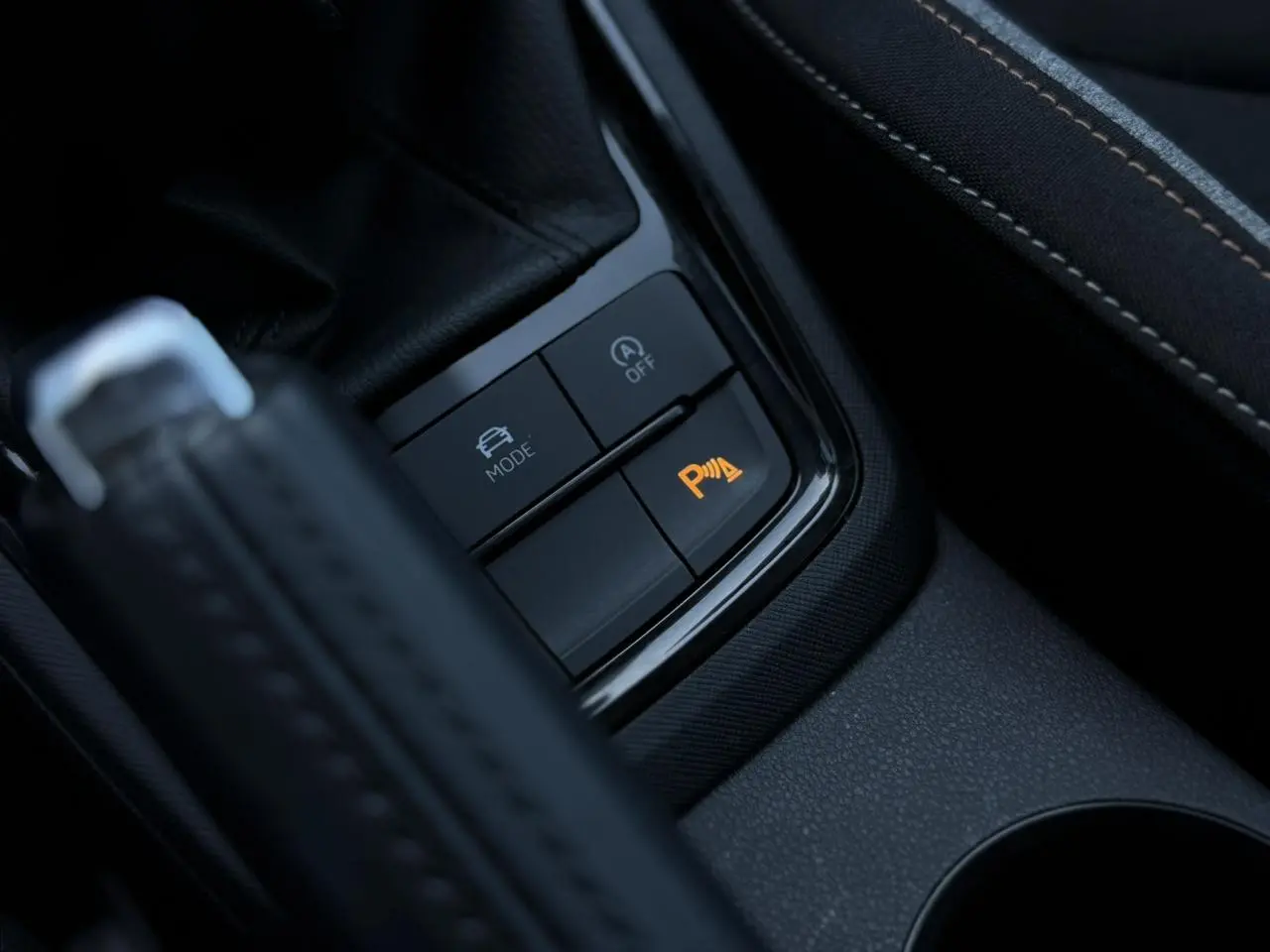 Gros plan sur les commandes de la console centrale avec frein à main et bouton orange d’aide au stationnement actif.