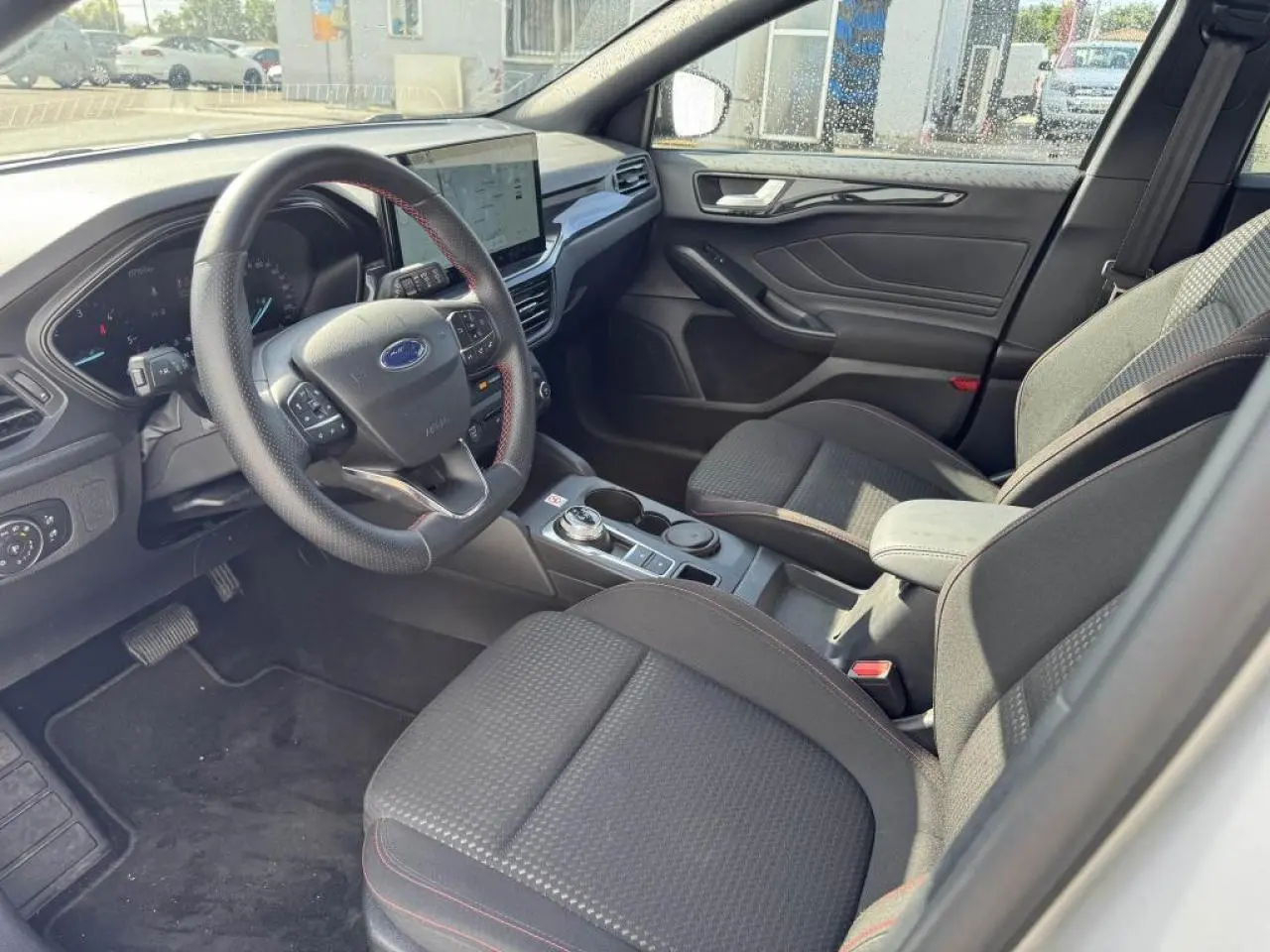 Vue intérieure côté conducteur de la Ford Focus IV ST-Line avec sellerie tissu noire et surpiqûres rouges.