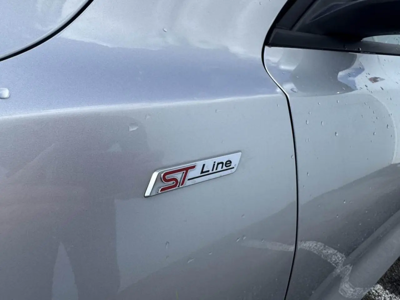 Gros plan sur le badge latéral ST-Line de la Ford Focus IV gris Moondust Metal, côté gauche avec gouttes d'eau.