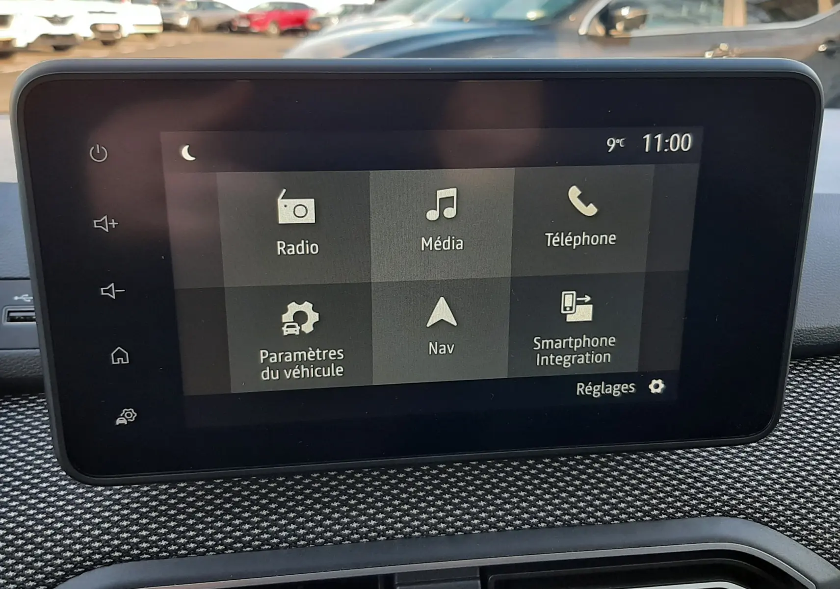Vue intérieure centrée sur l'écran tactile du tableau de bord du Dacia Jogger 2025, avec commandes visibles et arrière-plan extérieur flou.