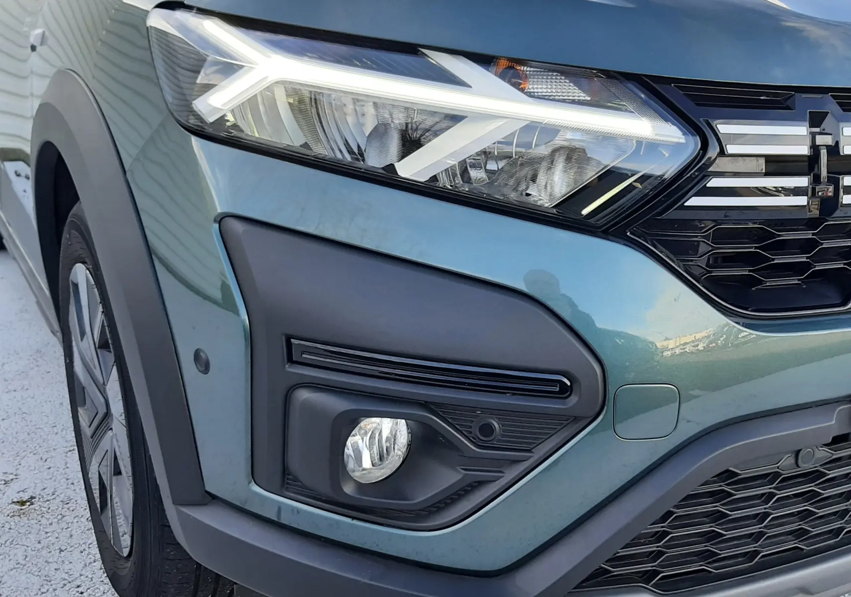 Vue 3/4 avant d'un Dacia Jogger 2025 vert cèdre métal avec toit noir et calandre distinctive, stationné devant un mur blanc.