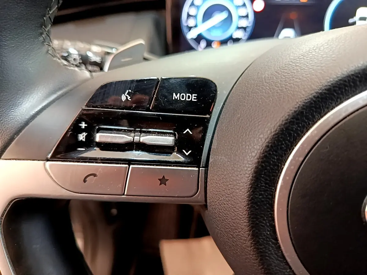 Gros plan sur les commandes gauche du volant cuir noir du Hyundai Tucson 1.6 Hybrid 230, tableau de bord numérique en arrière-plan.