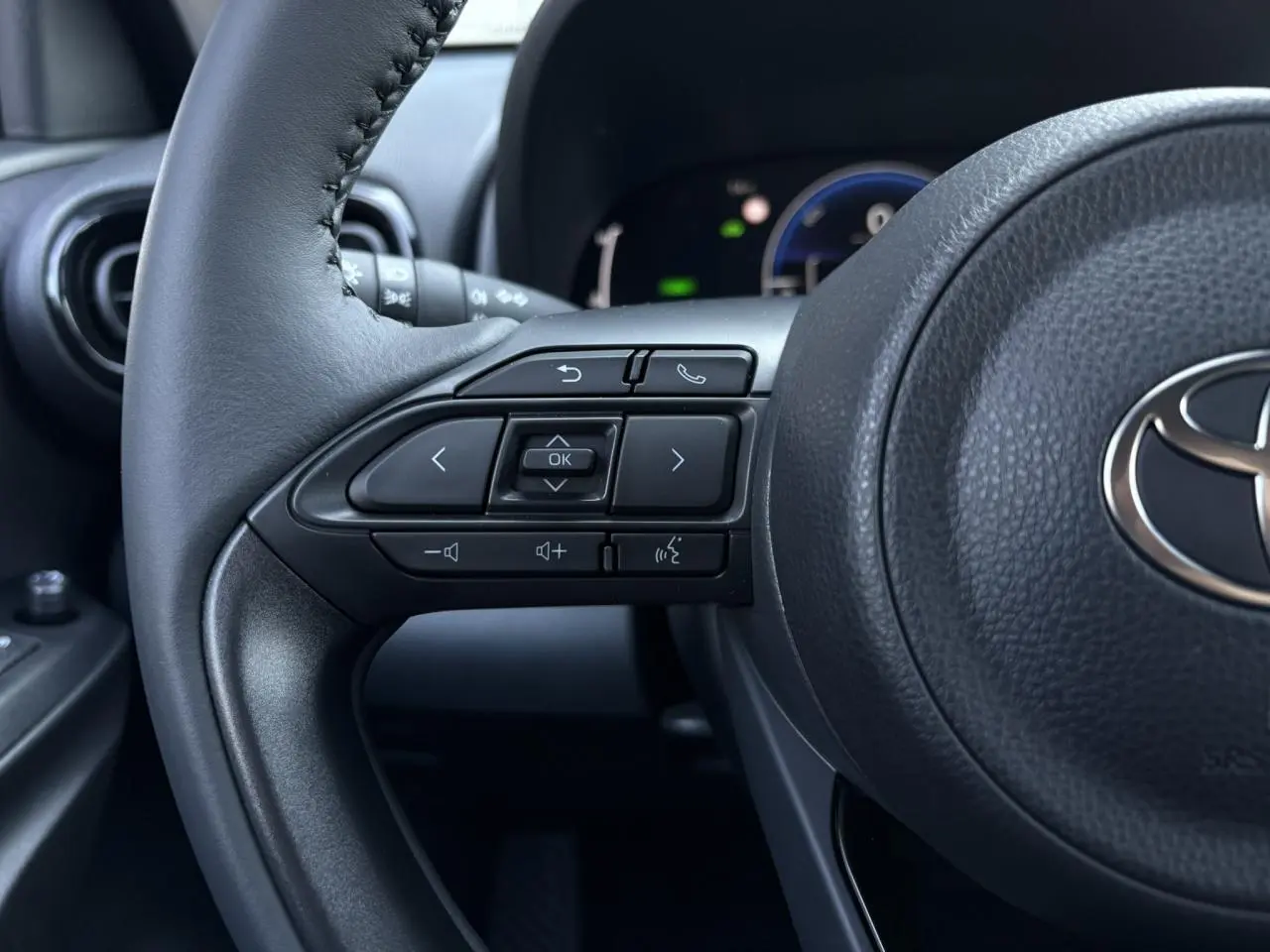 Gros plan sur le volant cuir noir de la Toyota Yaris 2025, avec commandes intégrées et tableau de bord numérique en arrière-plan.