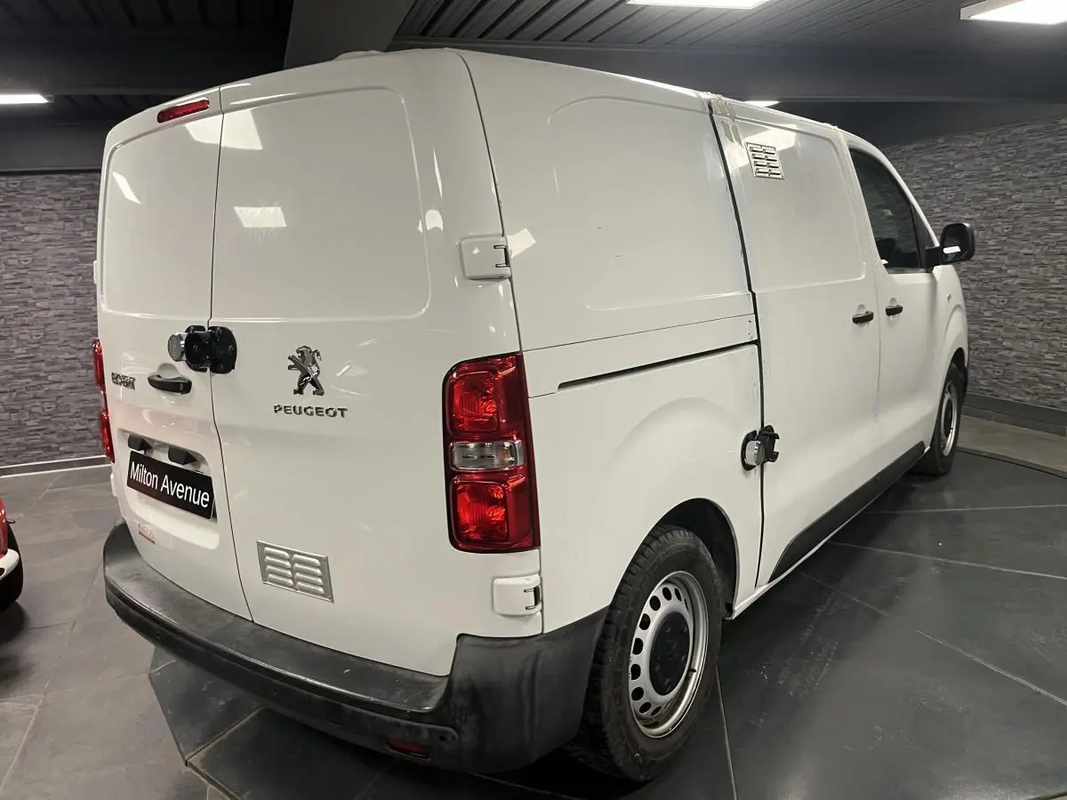 Vue 3/4 arrière droite d'un Peugeot Expert Fourgon blanc avec boucliers noirs et portes tôlées en intérieur showroom.
