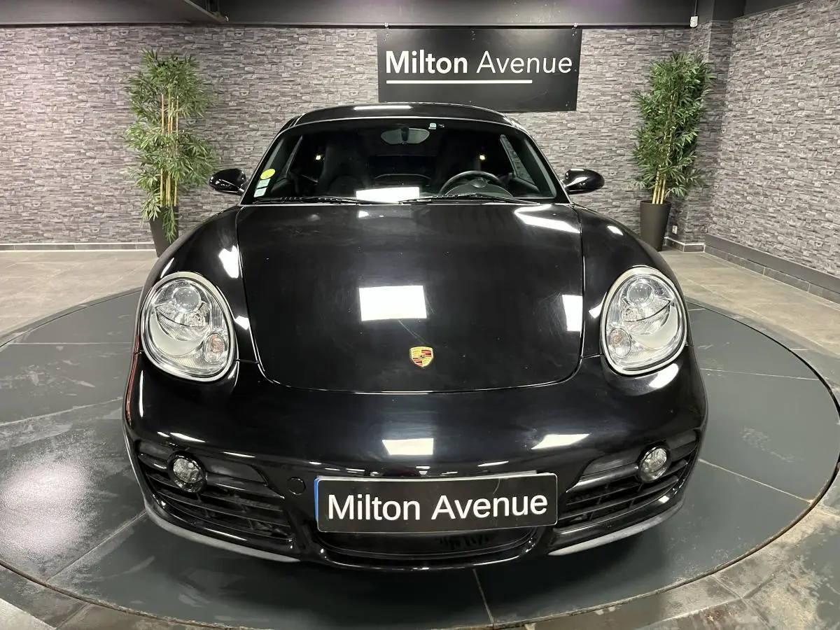Vue avant d'une Porsche Cayman noire 2008 avec phares ronds et plaque Milton Avenue en intérieur showroom.