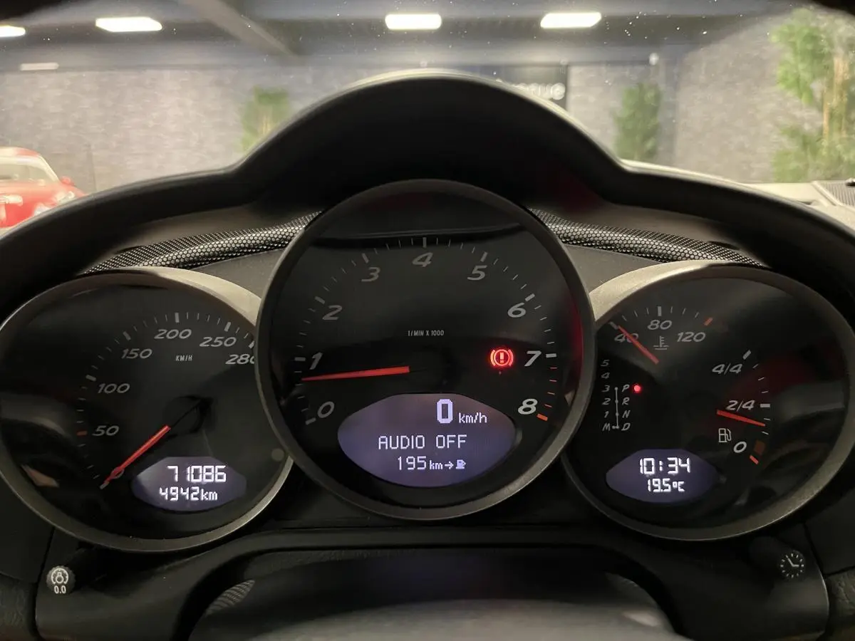 Gros plan sur le tableau de bord noir du Porsche Cayman 987 avec compte-tours central et affichages numériques.