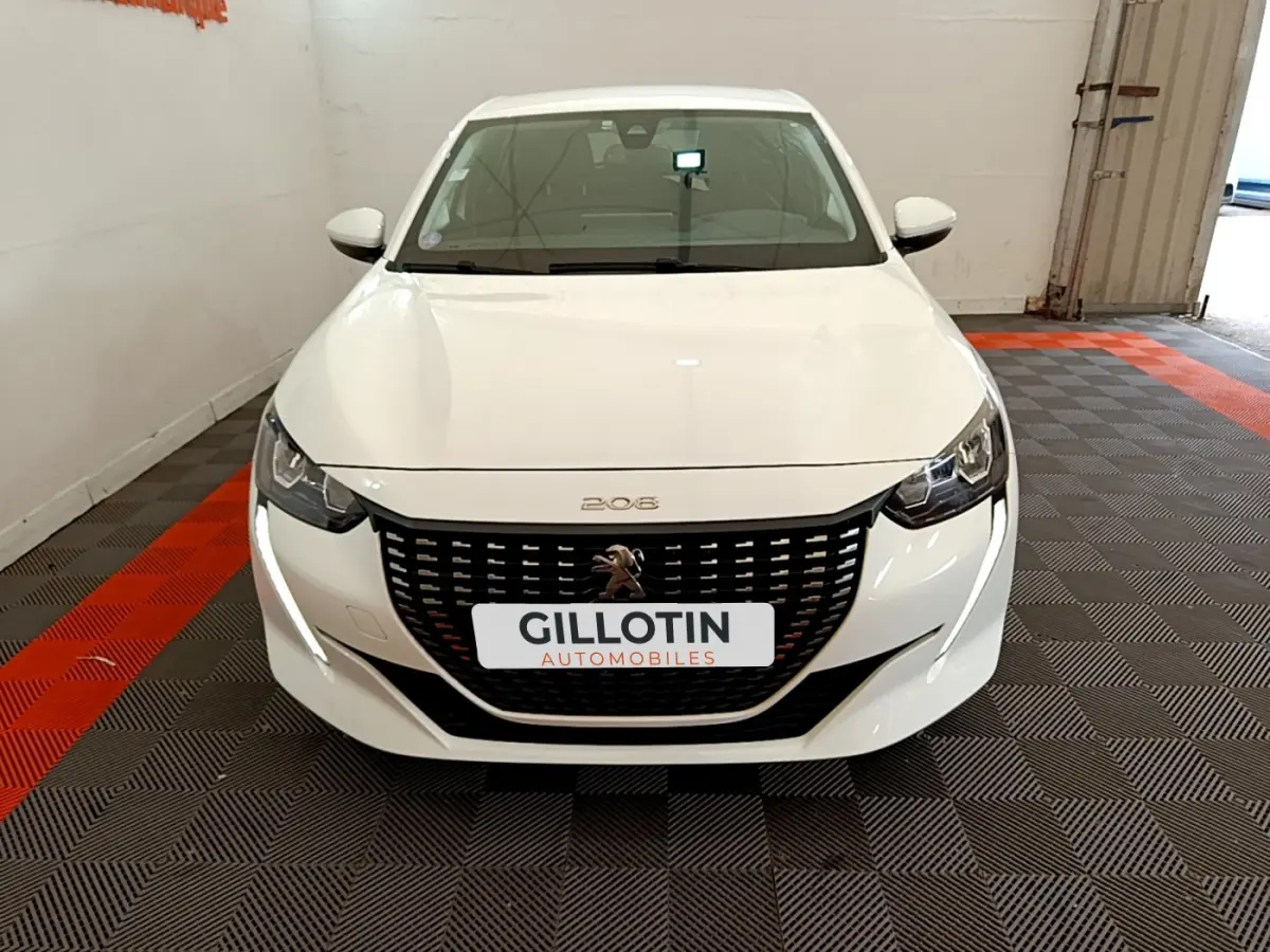 Vue avant d'une Peugeot 208 blanche 2020 avec calandre noire et feux LED, stationnée sur sol à damier gris et rouge.