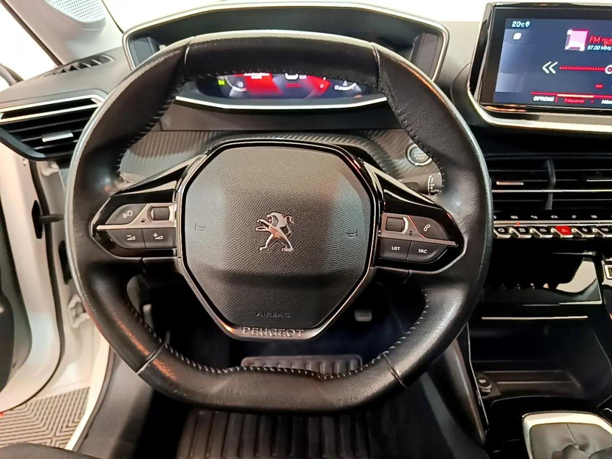 Vue rapprochée du volant cuir noir de la Peugeot 208 blanche, avec tableau de bord digital et écran tactile central visible.