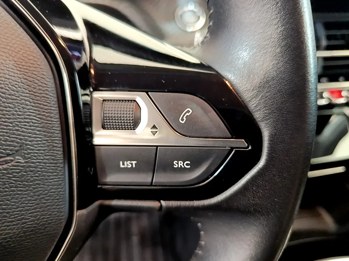 Gros plan sur les commandes du volant côté gauche en cuir noir du Peugeot 208 blanc PureTech 100 S&S Allure Business.